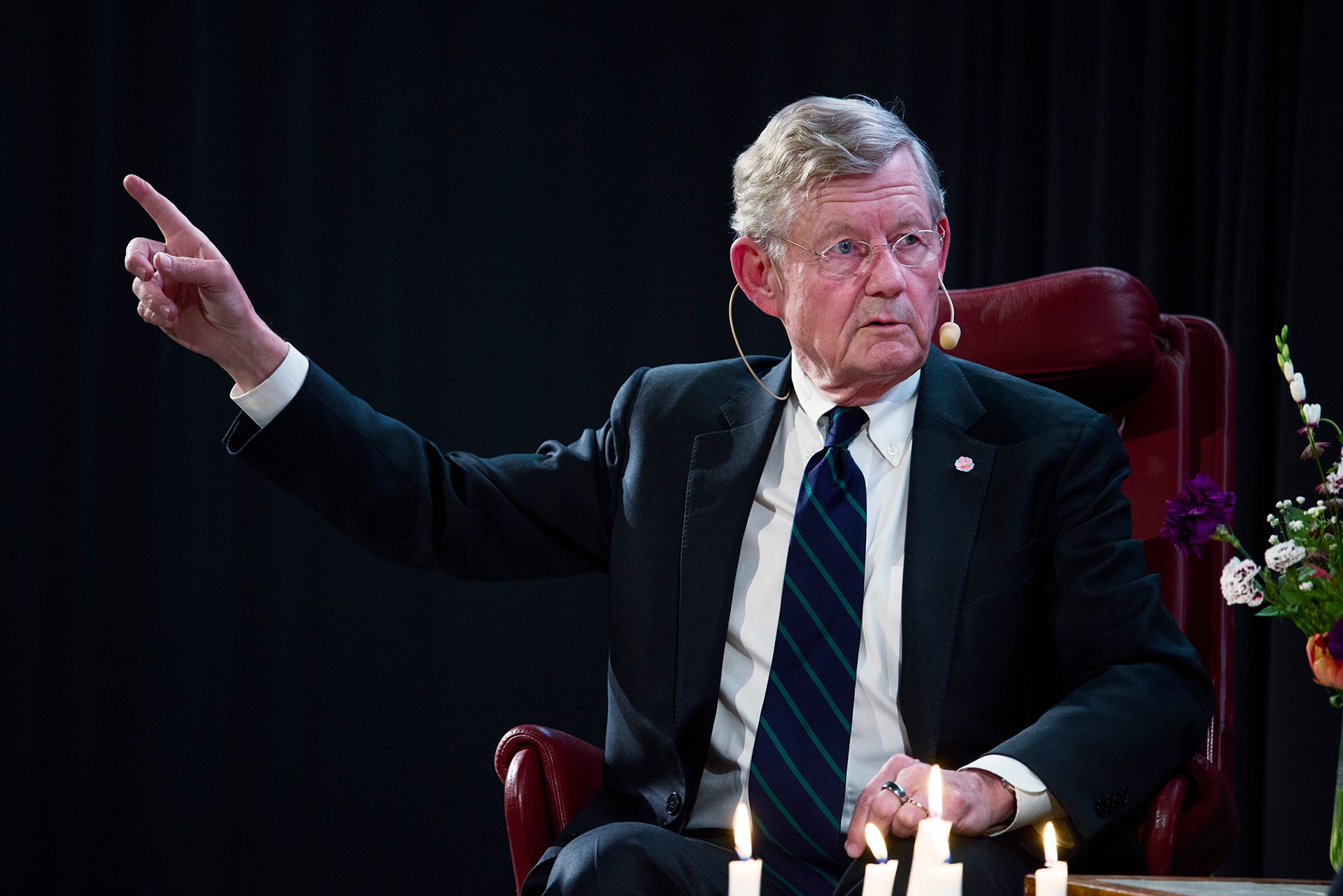 En äldre man i mörk kostym och slips sitter i en röd stol och gestikulerar medan han talar mot en mörk bakgrund.