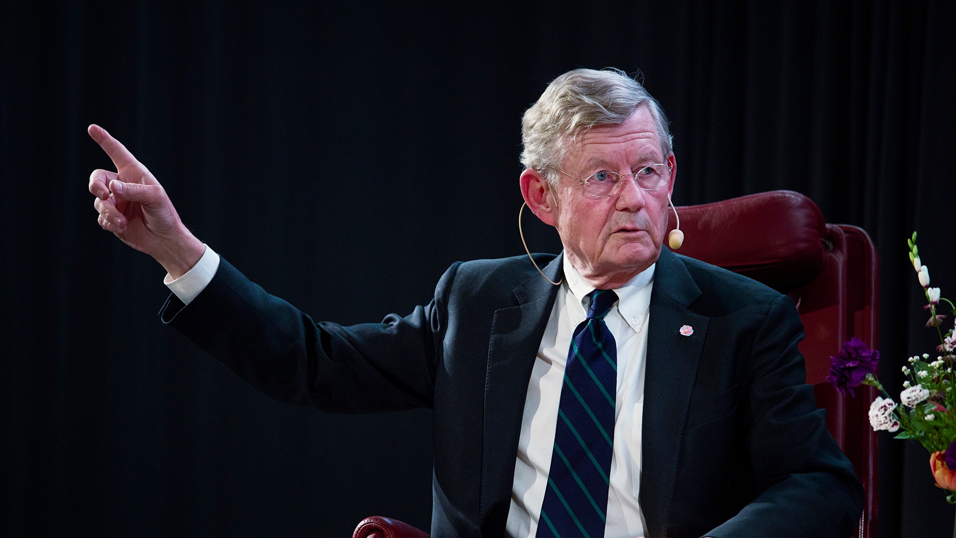 En äldre man i mörk kostym och slips sitter i en röd stol och gestikulerar medan han talar mot en mörk bakgrund.