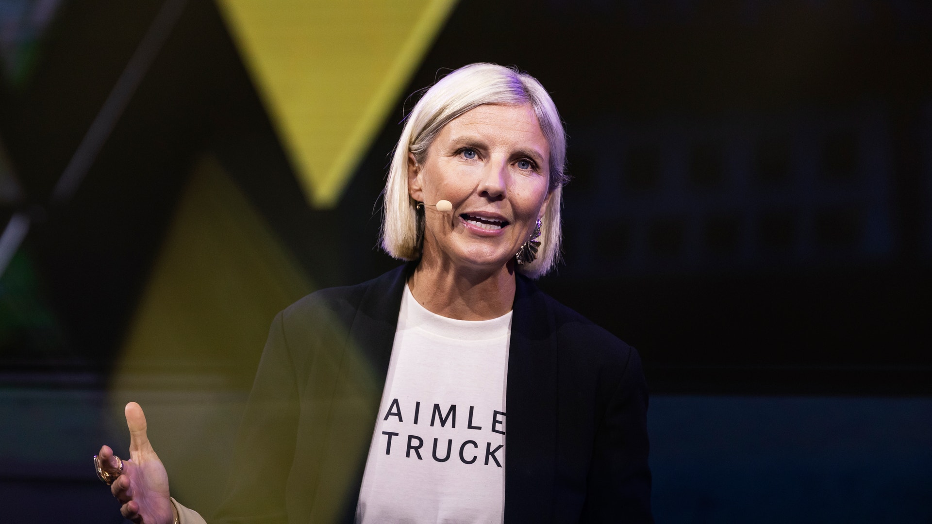 En kvinna talar på scen med hjälp av en headset-mikrofon och gestikulerar med handen.