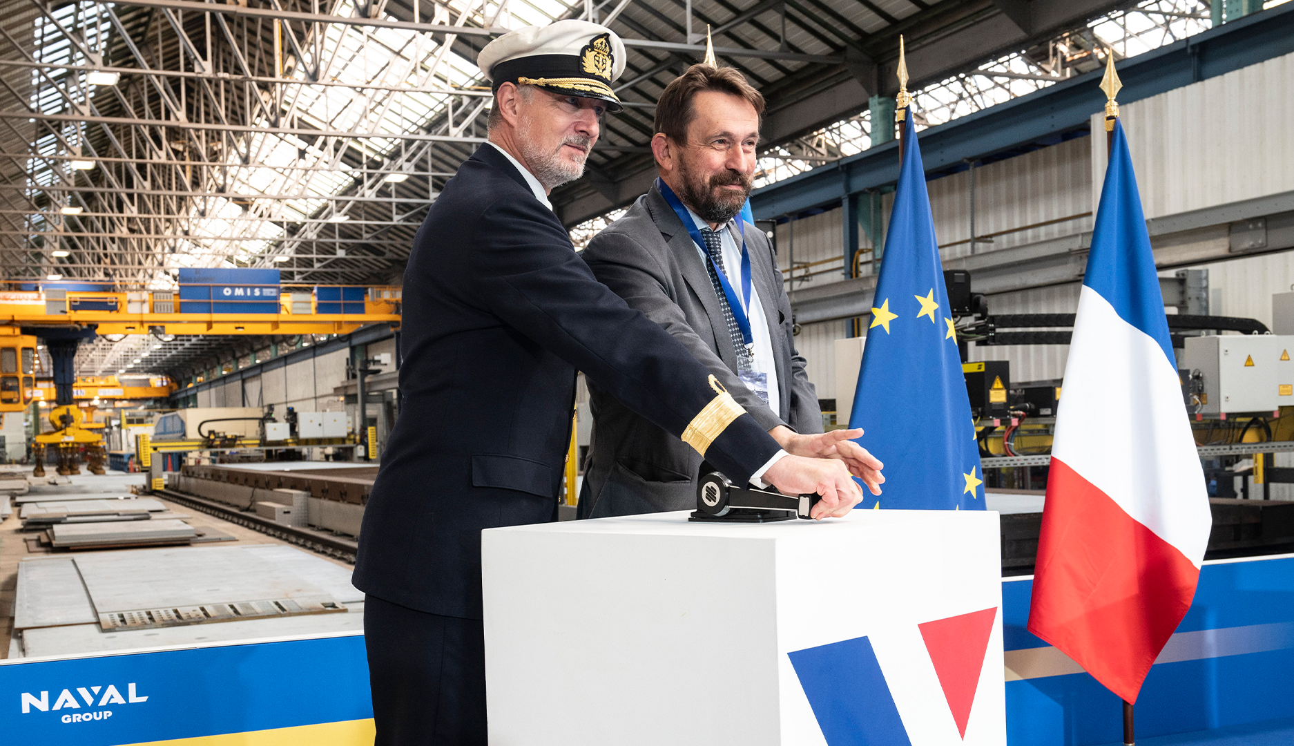 En marinofficer och en man i kostym står vid en ceremoni i en industrihall, med EU-flaggan och franska flaggan samt Naval Groups logotyp i bakgrunden.