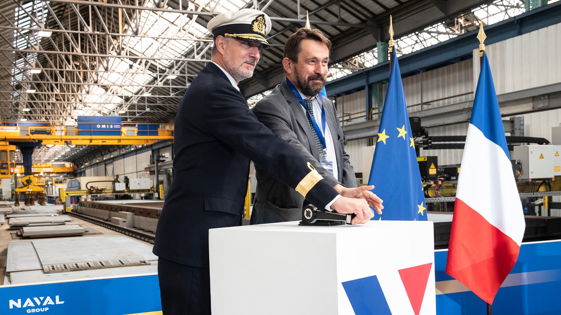 En marinofficer och en man i kostym står vid en ceremoni i en industrihall, med EU-flaggan och franska flaggan samt Naval Groups logotyp i bakgrunden.