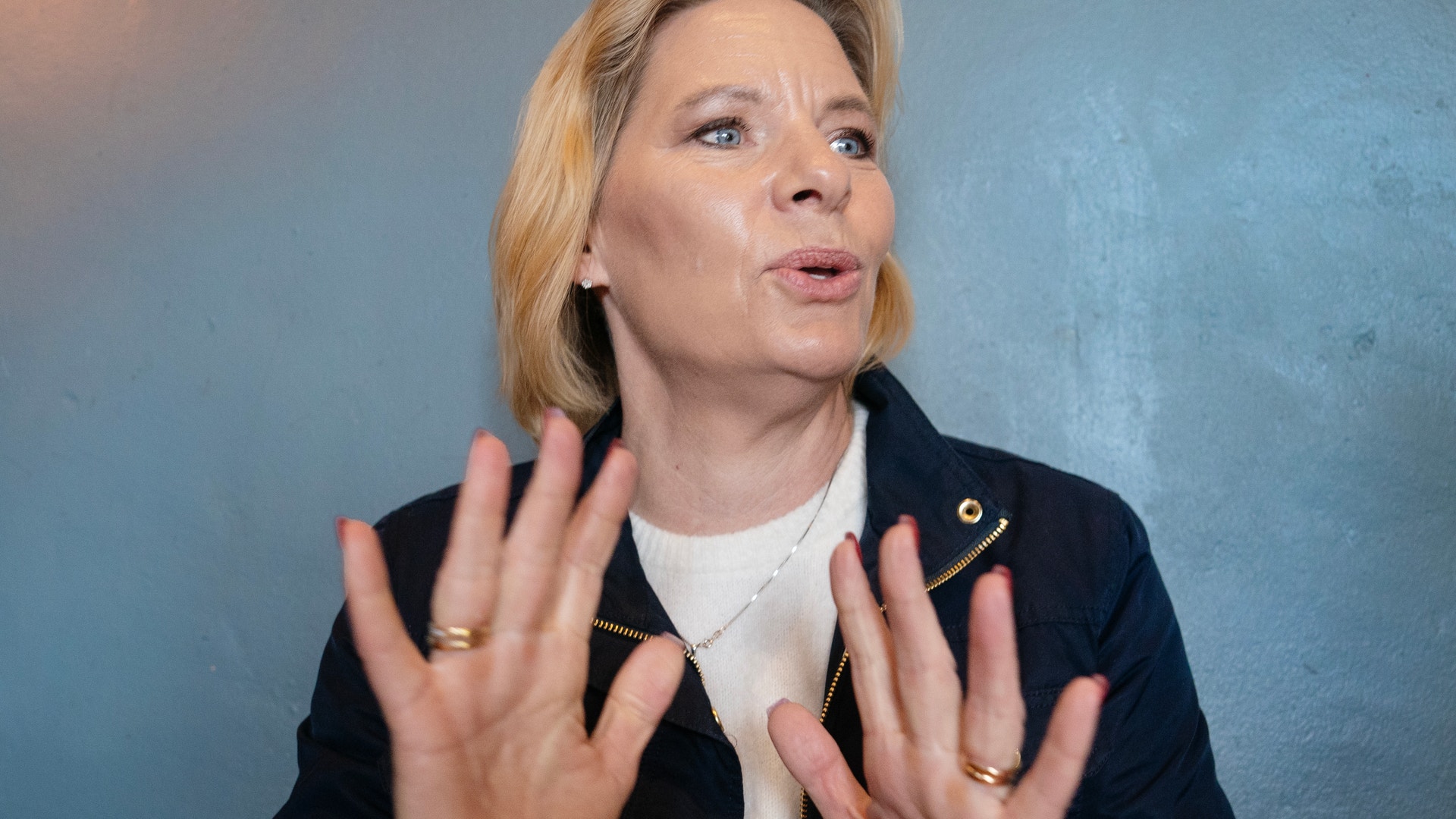 Kvinna med blont hår gestikulerar med båda händerna. Hon bär en svart blazer och vit tröja mot en blå bakgrund.