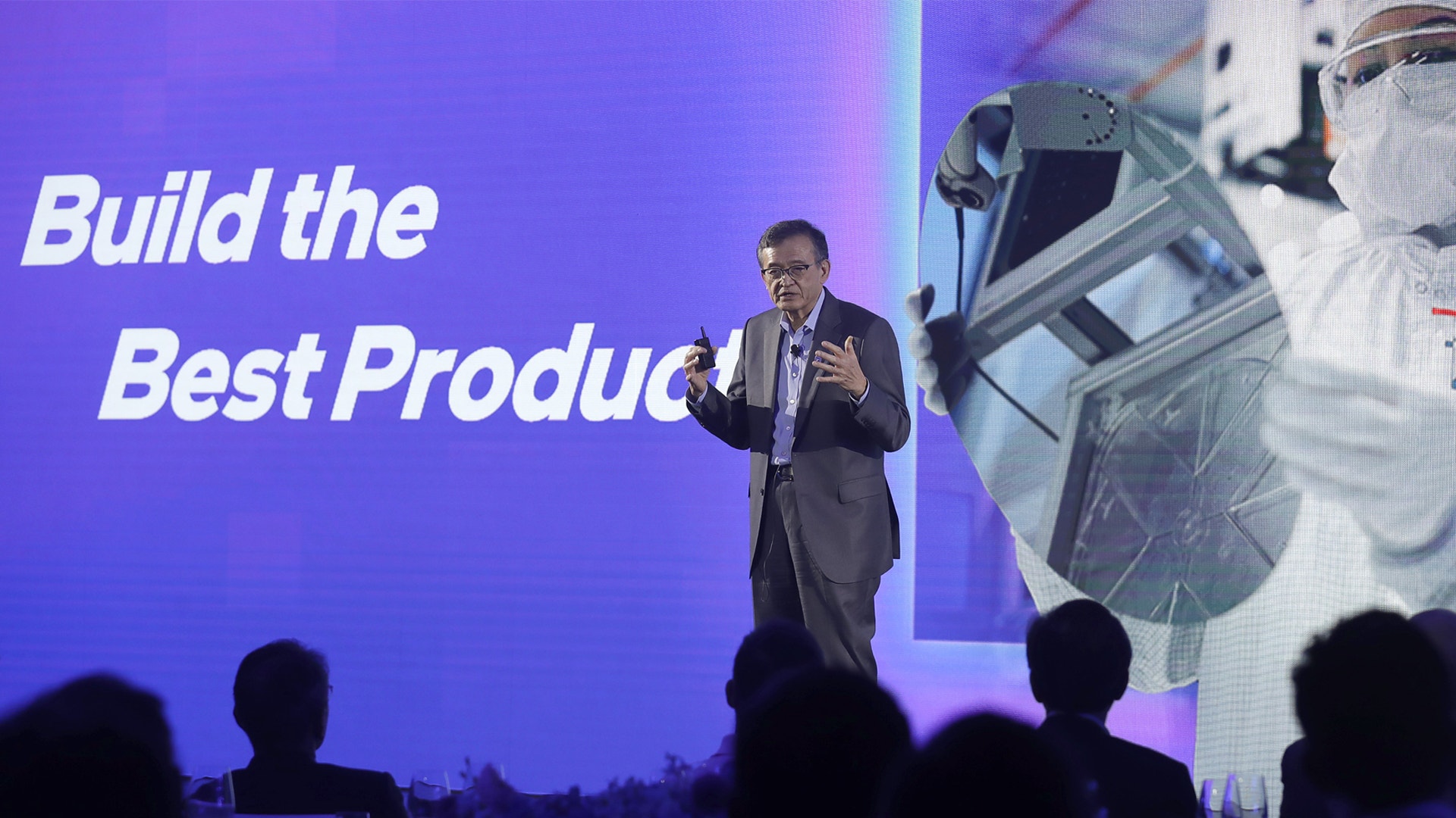 En man i mörk kostym presenterar på en scen med lila bakgrund och texten 'Build the Best Product'. Publik syns i förgrunden.