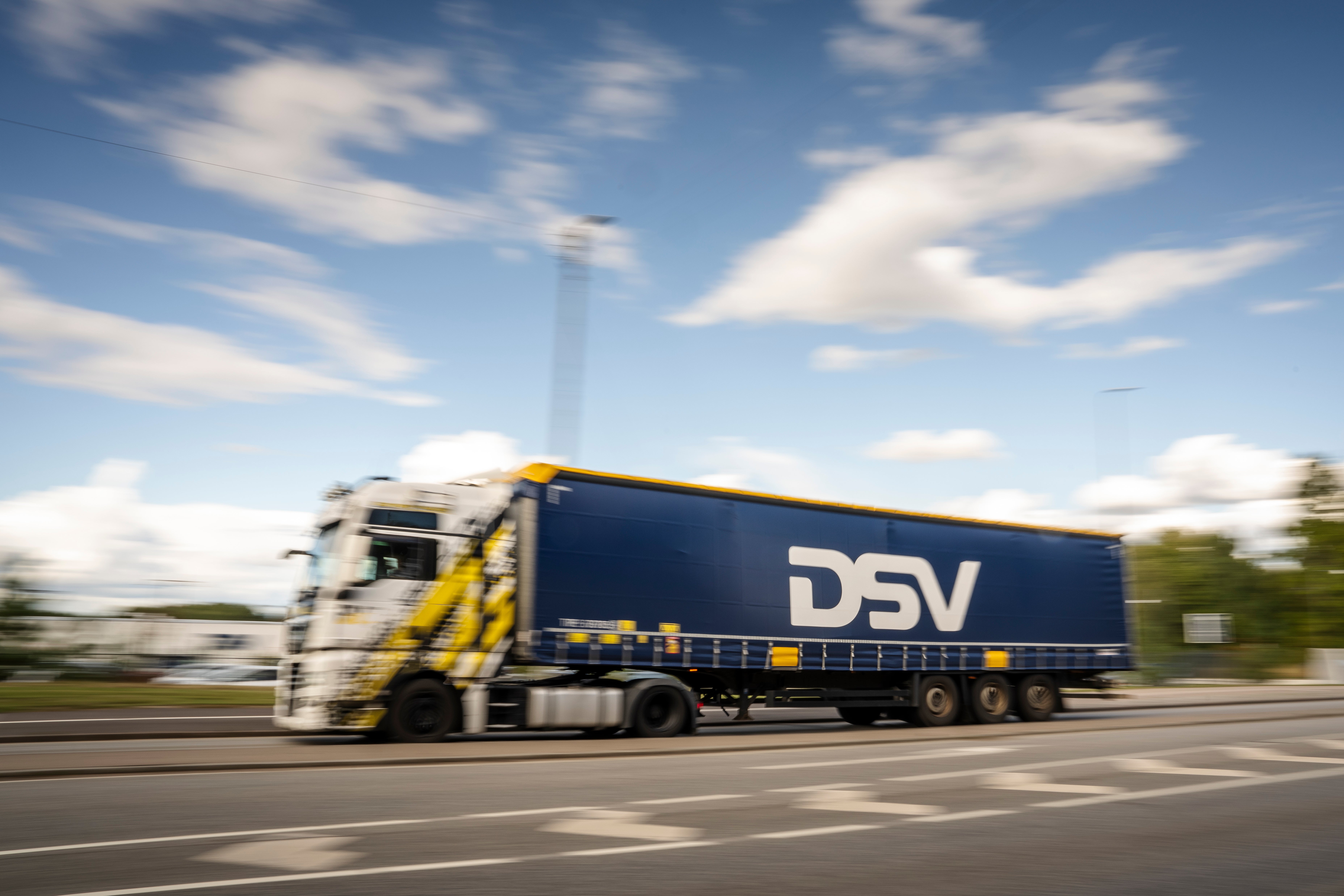 En blå och gul lastbil med DSV-logotyp kör på en motorväg under en solig himmel med moln.