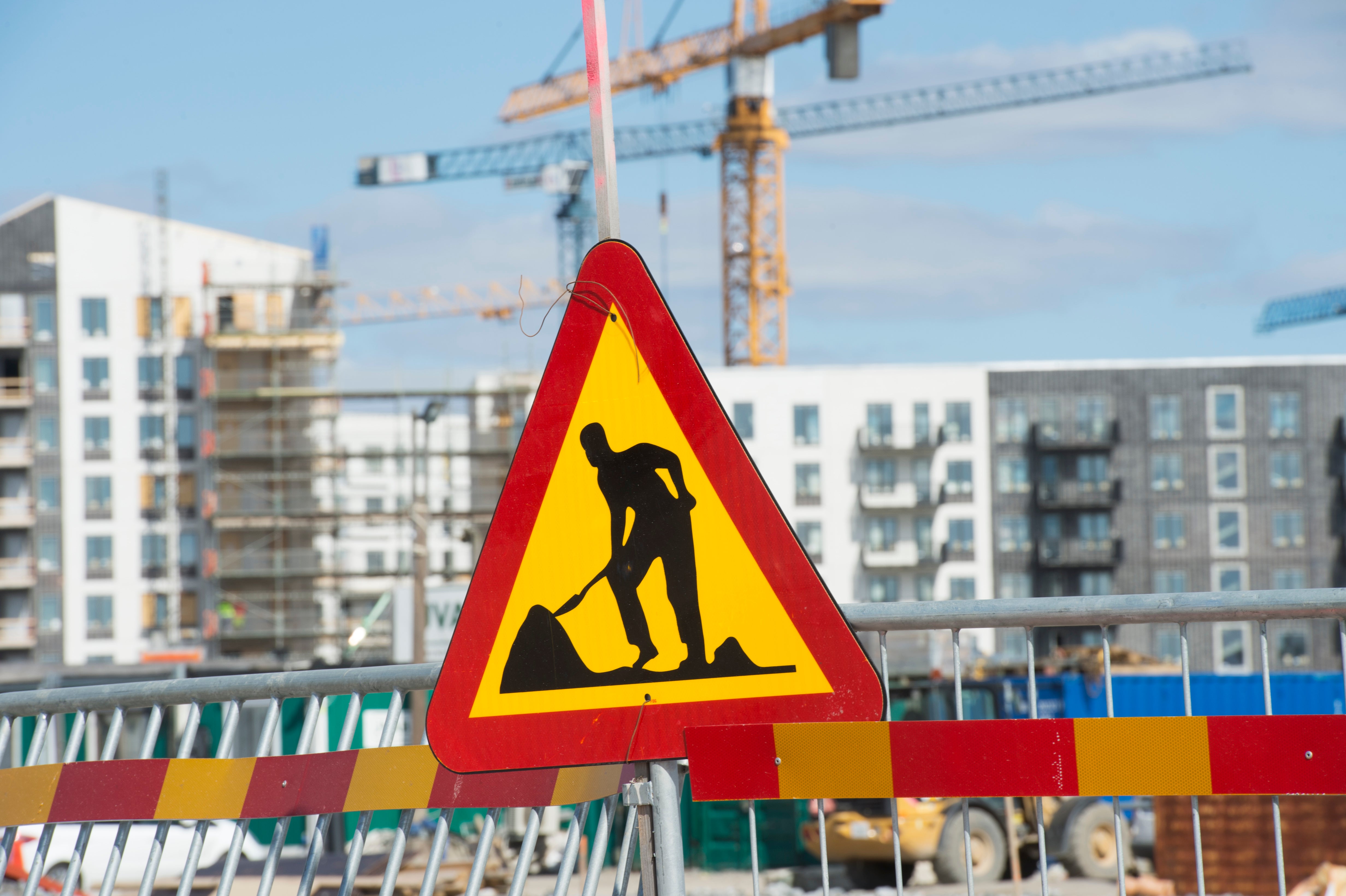 Varningsskylt för pågående konstruktion på en byggarbetsplats med höga hus och byggkranar i bakgrunden.