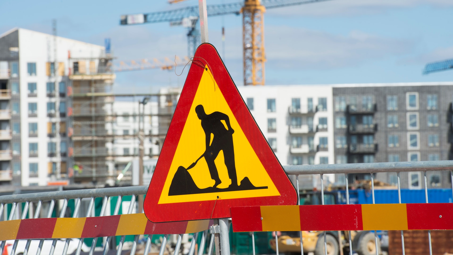 Varningsskylt för pågående konstruktion på en byggarbetsplats med höga hus och byggkranar i bakgrunden.