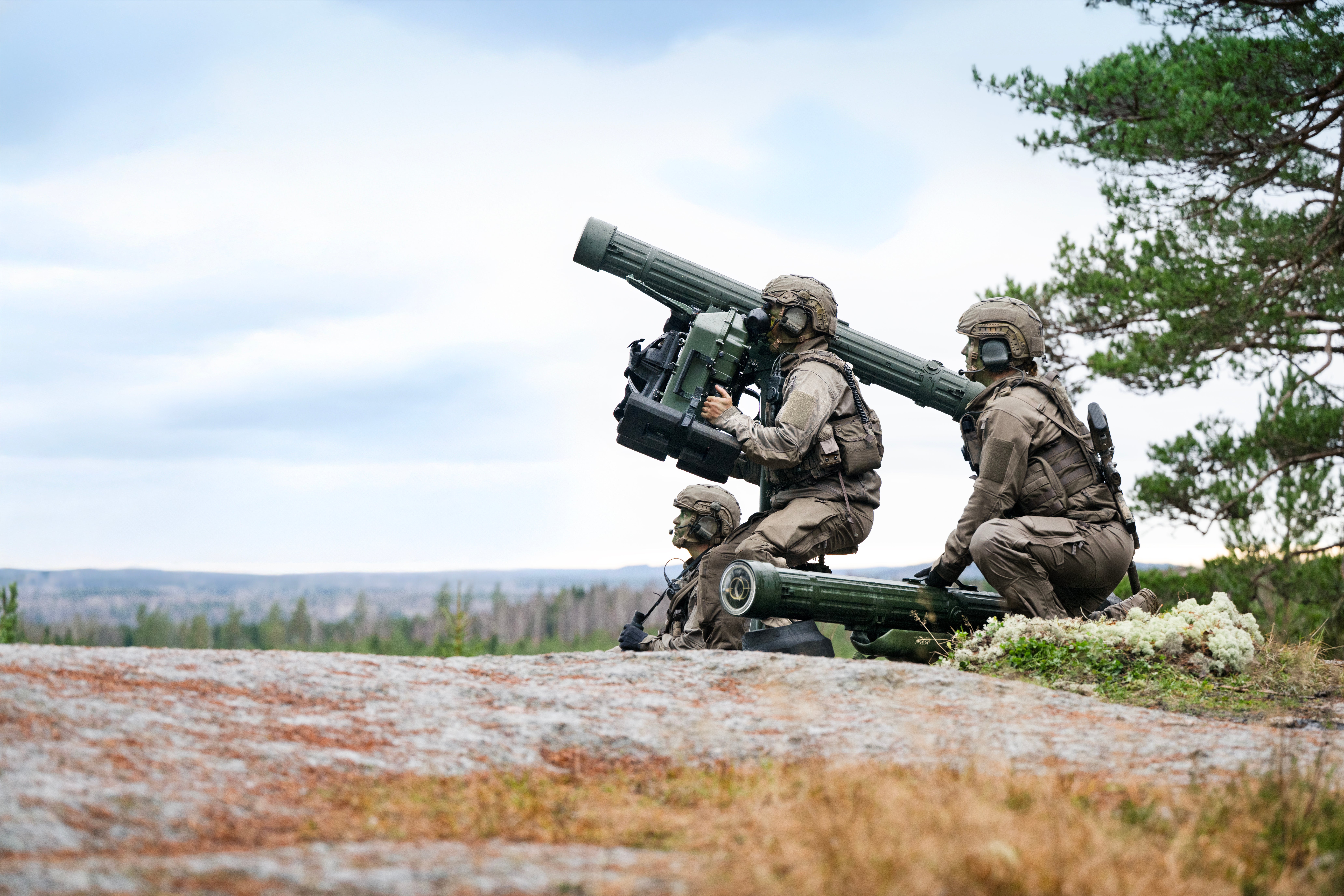 Två soldater i militär utrustning använder ett portabelt robotvapen i öppen terräng med skog i bakgrunden.