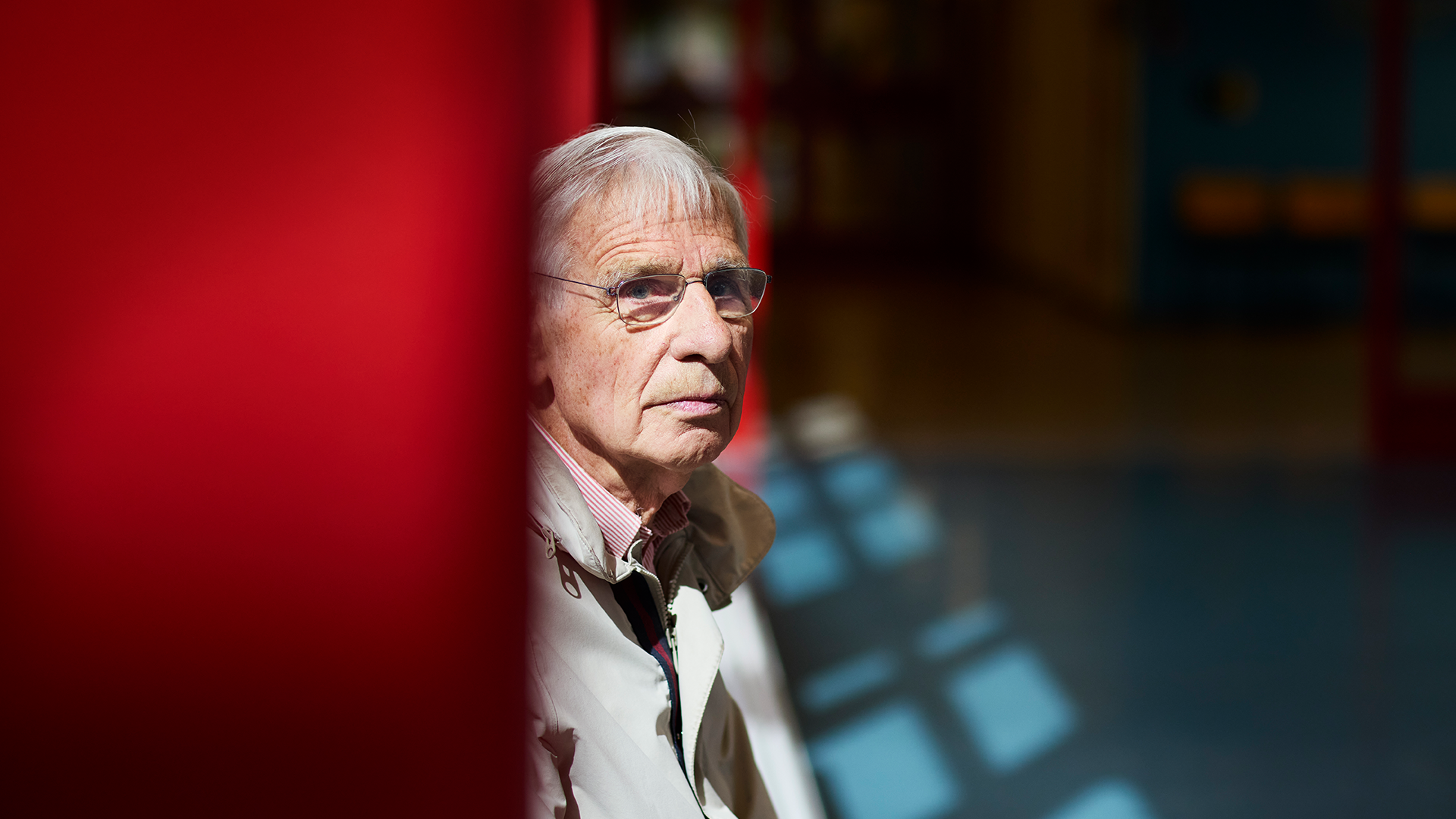 Porträttbild av en äldre man med glasögon, fotograferad mot röd bakgrund.