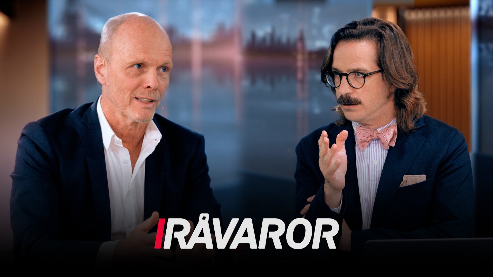 Två män i professionell miljö sitter och diskuterar framför en bakgrund med Londons skyline. Texten 'RÅVAROR' visas längst ned på bilden.