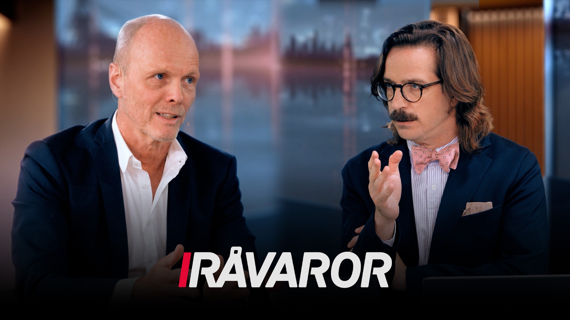 Två män i professionell miljö sitter och diskuterar framför en bakgrund med Londons skyline. Texten 'RÅVAROR' visas längst ned på bilden.