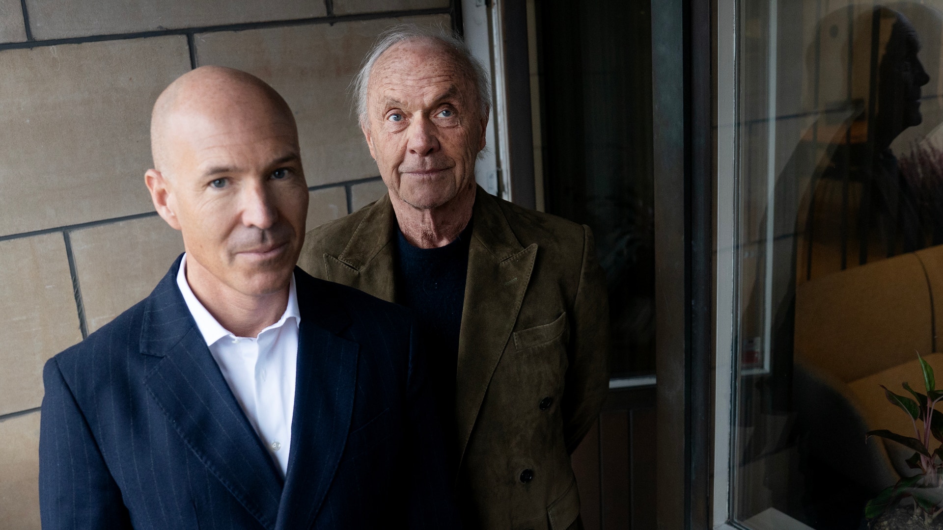 Två män står tillsammans framför en vägg med moderna kakel och ett fönster.