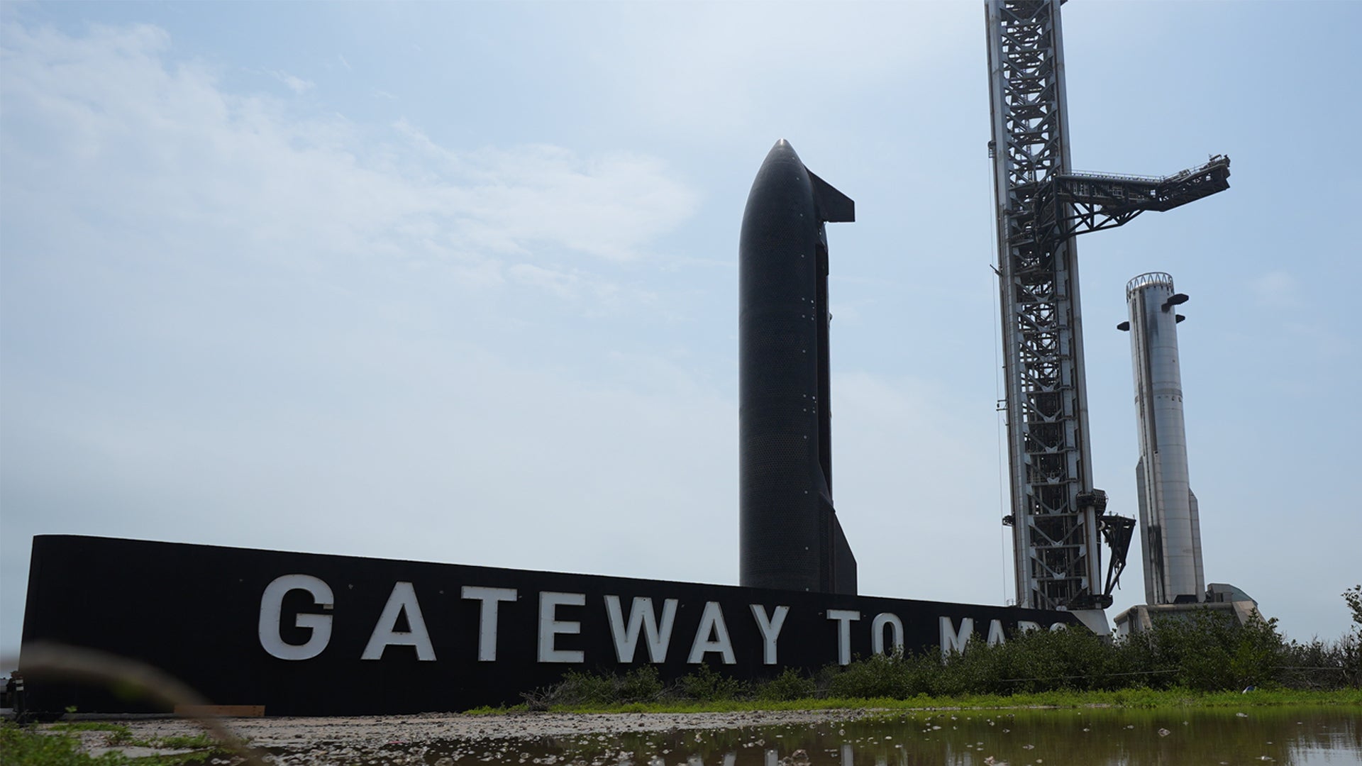 En stor svart metallskulptur av en raket med texten "Gateway to Mars" framför. Industriella kranar och konstruktioner syns i bakgrunden mot en molnig himmel.