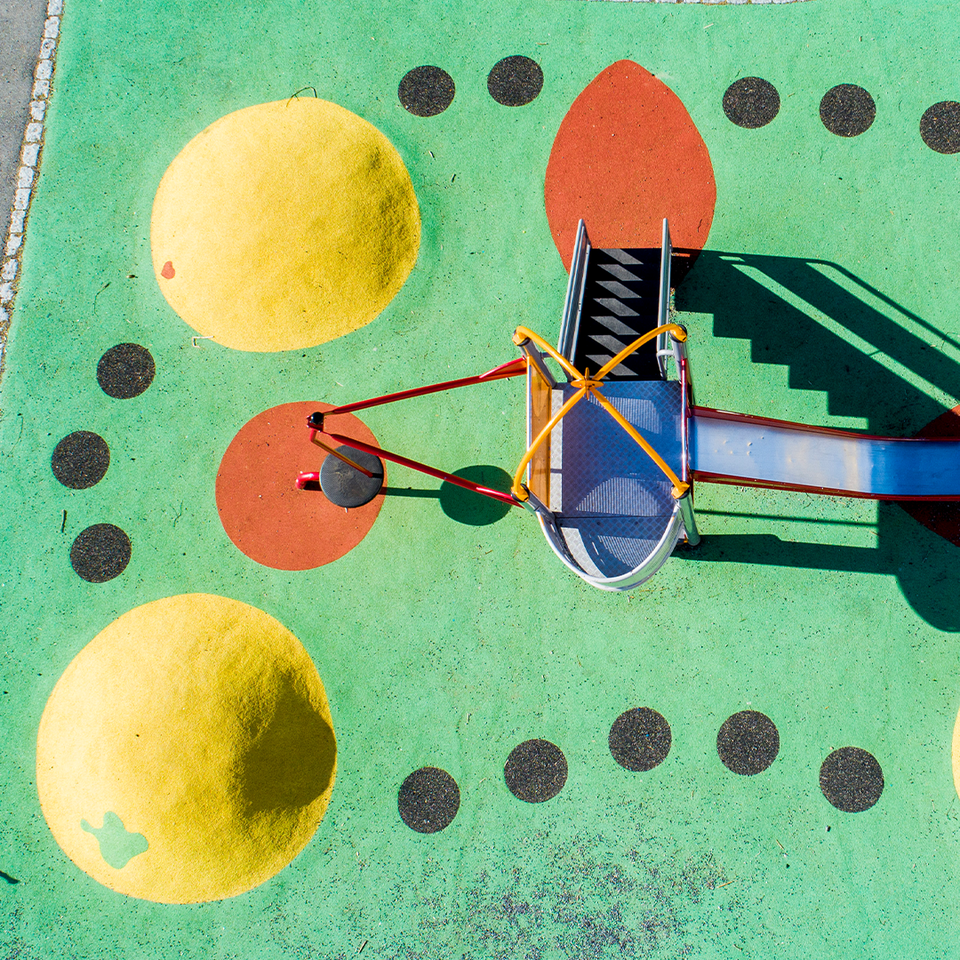 En färgglad lekplatsinstallation sedd från ovan med en grön yta. I mitten finns en abstrakt skulptur gjord av färgade metallrör i blått, rött, gult och grönt som bildar geometriska former. Runt skulpturen är det arrangerat gula bollar och mörkgrå cirklar i ett mönster. Till höger syns en gul och blå yta som är del av lekplatsen. Installationen är designad för att stimulera kreativitet och lek hos barn.