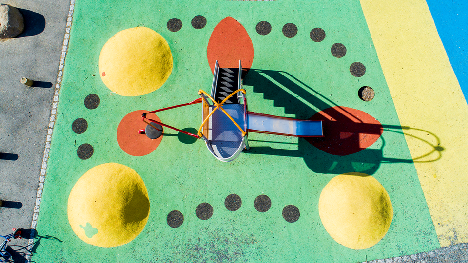 En färgglad lekplatsinstallation sedd från ovan med en grön yta. I mitten finns en abstrakt skulptur gjord av färgade metallrör i blått, rött, gult och grönt som bildar geometriska former. Runt skulpturen är det arrangerat gula bollar och mörkgrå cirklar i ett mönster. Till höger syns en gul och blå yta som är del av lekplatsen. Installationen är designad för att stimulera kreativitet och lek hos barn.