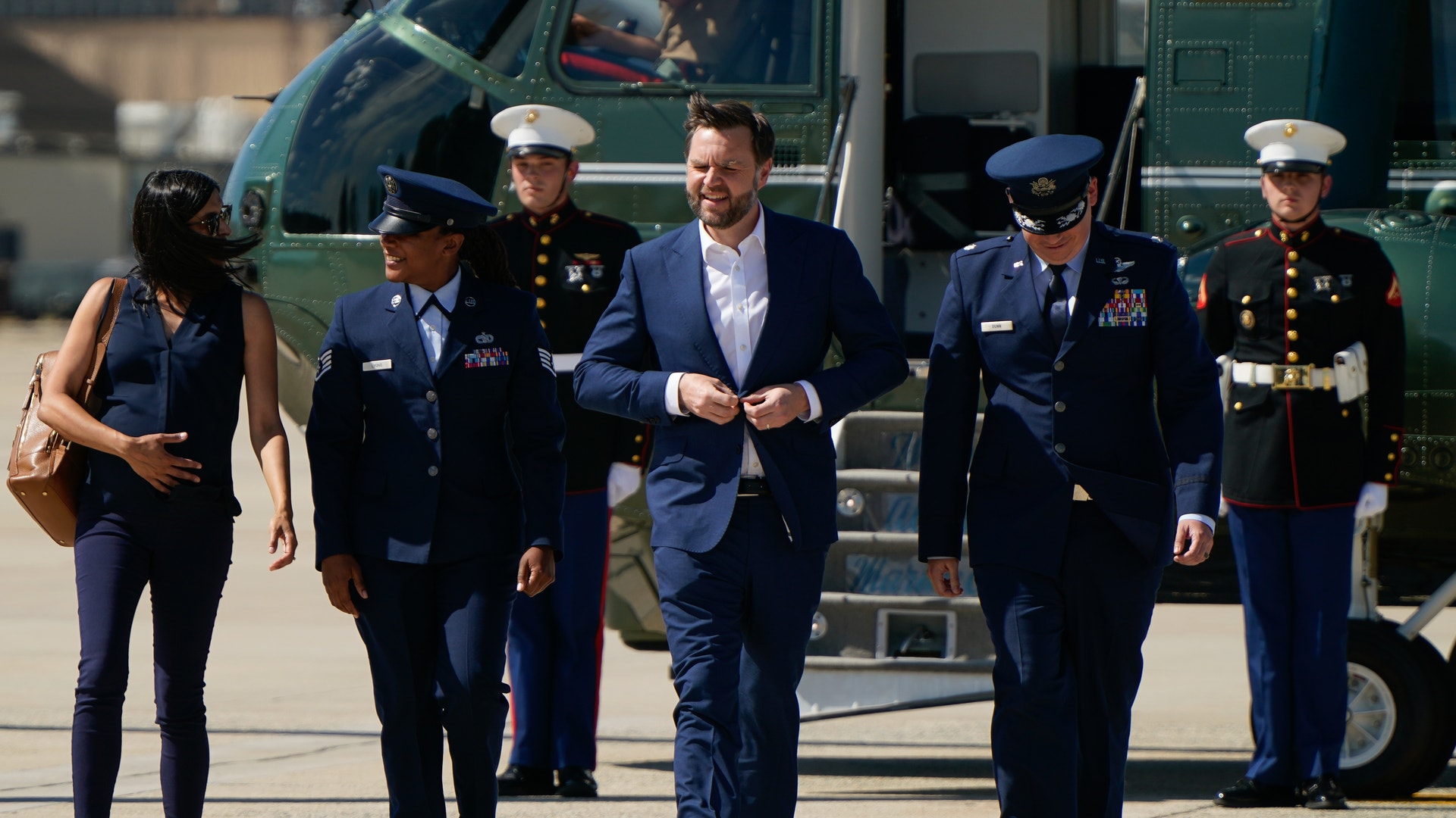 En man i blå kostym går tillsammans med militärer i uniformer på en flygplats. Ett militärflygplan syns i bakgrunden.