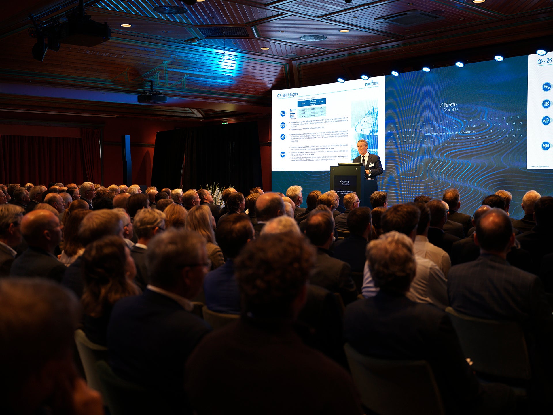 En person håller en presentation vid en talarstol inför en stor publik i en konferenssal, med en skärm som visar diagram i bakgrunden.