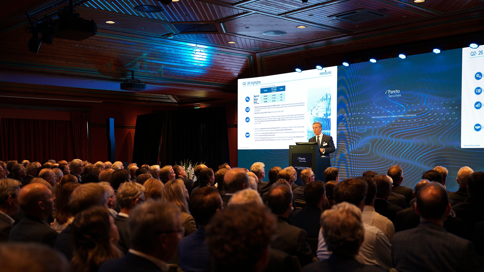 En person håller en presentation vid en talarstol inför en stor publik i en konferenssal, med en skärm som visar diagram i bakgrunden.