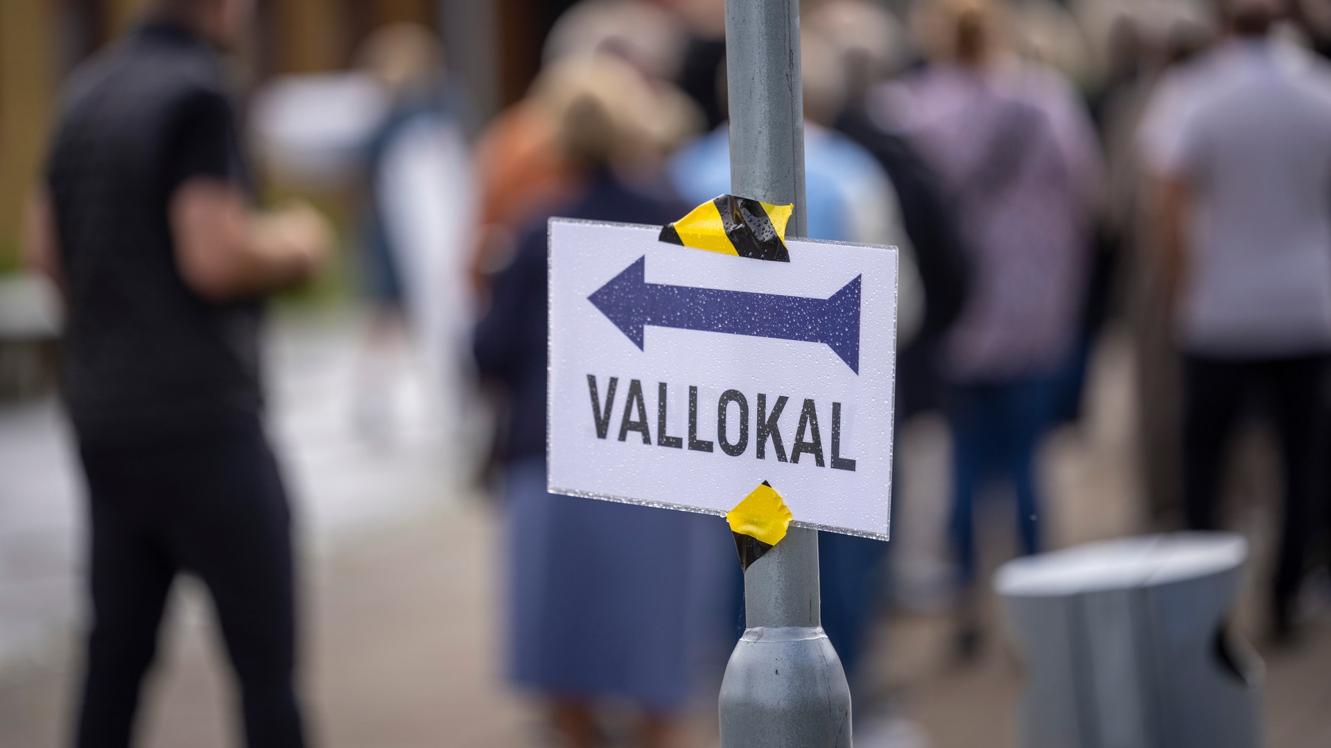 En skylt med texten "Vallokal" och en pil, med kö av människor i bakgrunden.