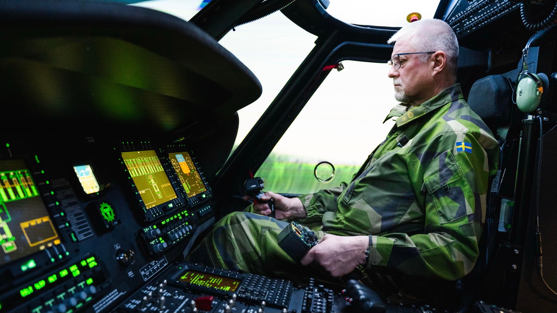 Militär pilot i helikopterkockpit med instrumentbräde och kontroller.
