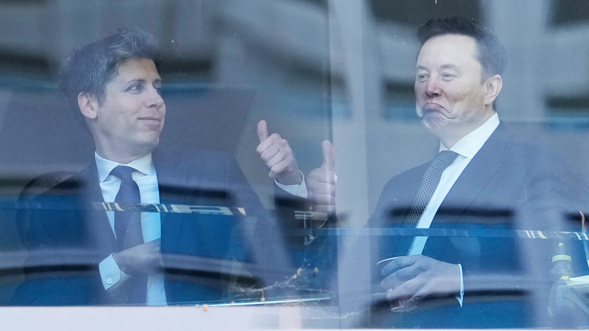 Två män i kostym diskuterar bakom ett glasfönster. Mannen till vänster gestikulerar medan han talar, mannen till höger lyssnar.