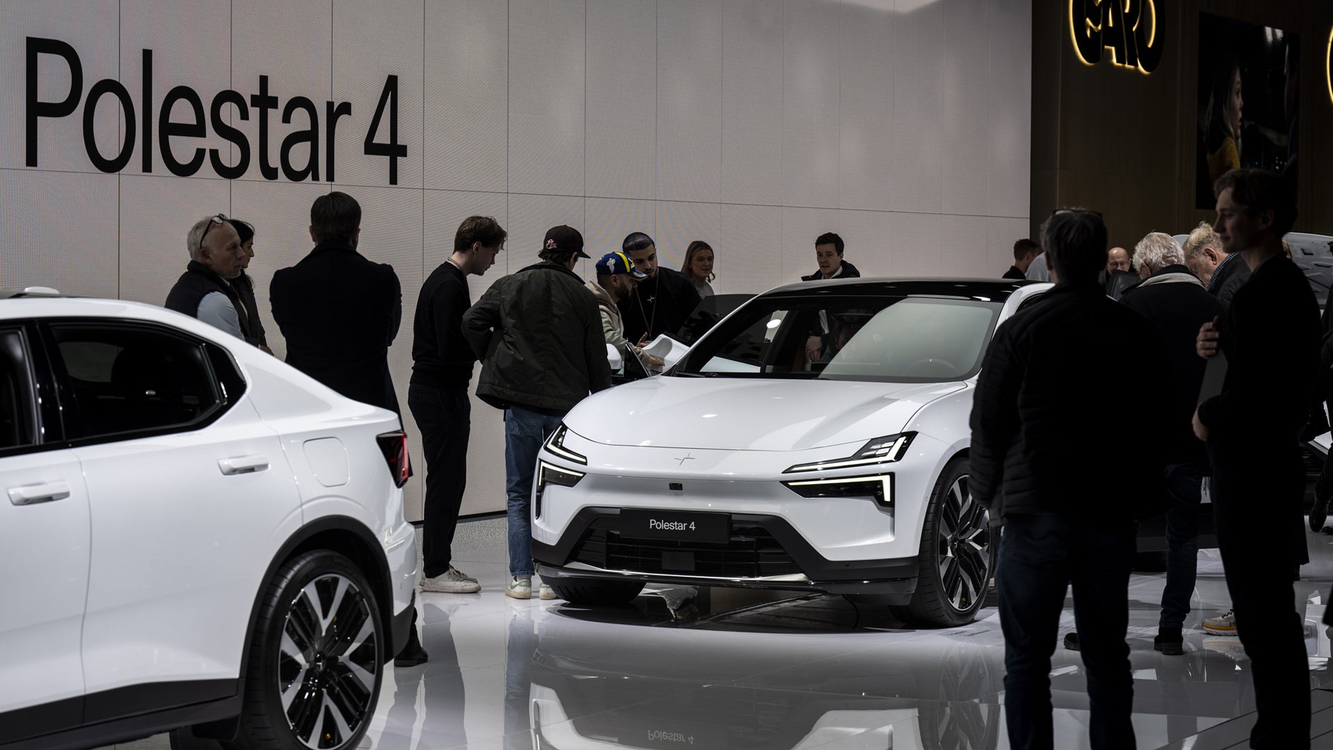 En vit Polestar 4 elfordon visas upp på en bilmässa. Flera besökare i mörka kläder står omkring bilen för att se den närmare. I bakgrunden syns texten Polestar 4 på en vägg.
