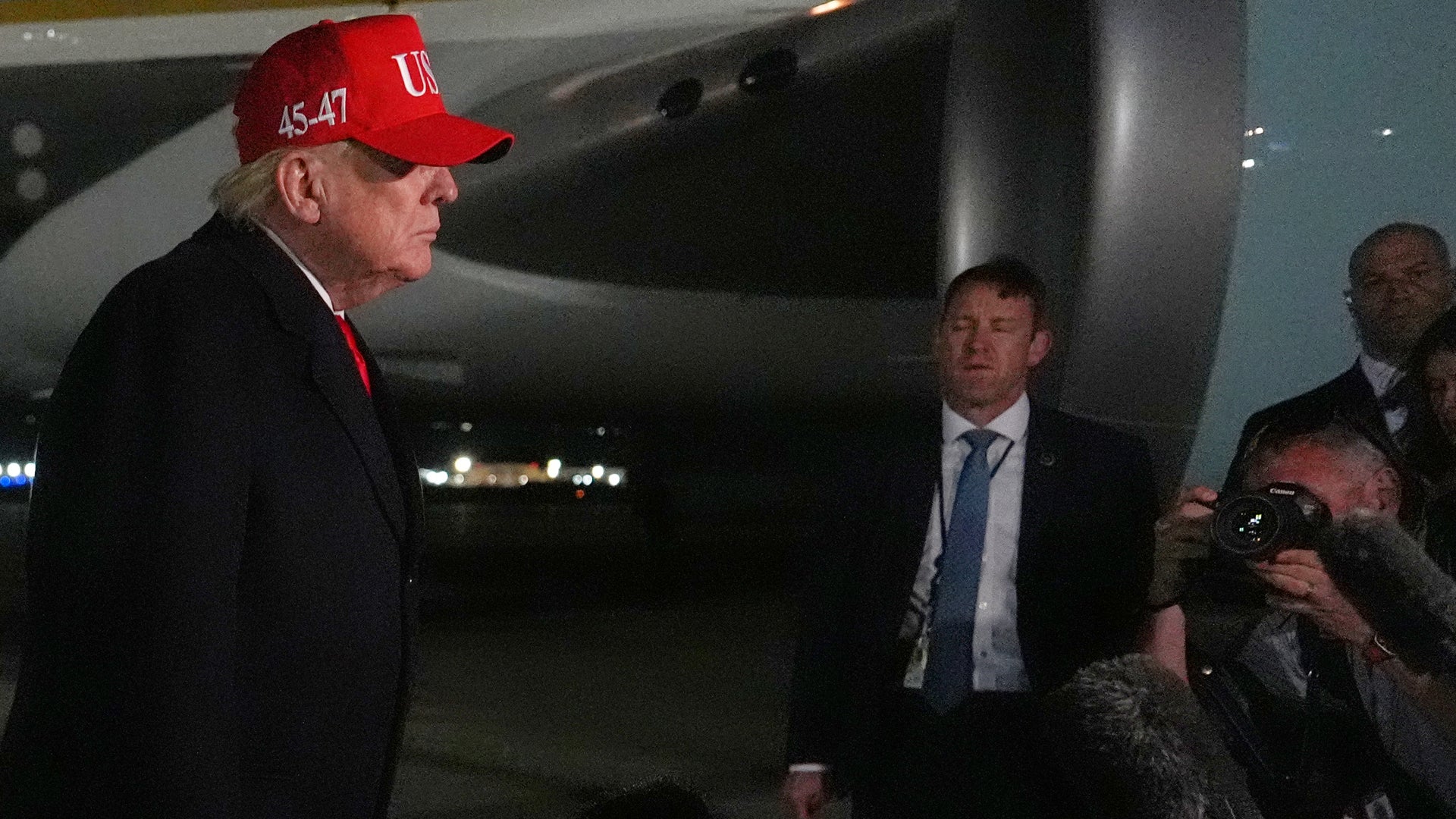 Man i röd keps och mörk kostym talar med flera personer inomhus med flygplan i bakgrunden.