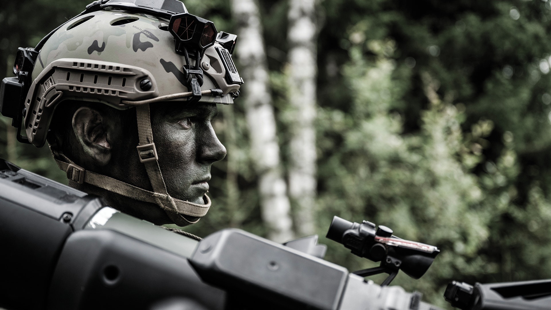 Soldat i militär utrustning med taktisk hjälm och gevär, fotograferad i profil mot en skogsbakgrund.