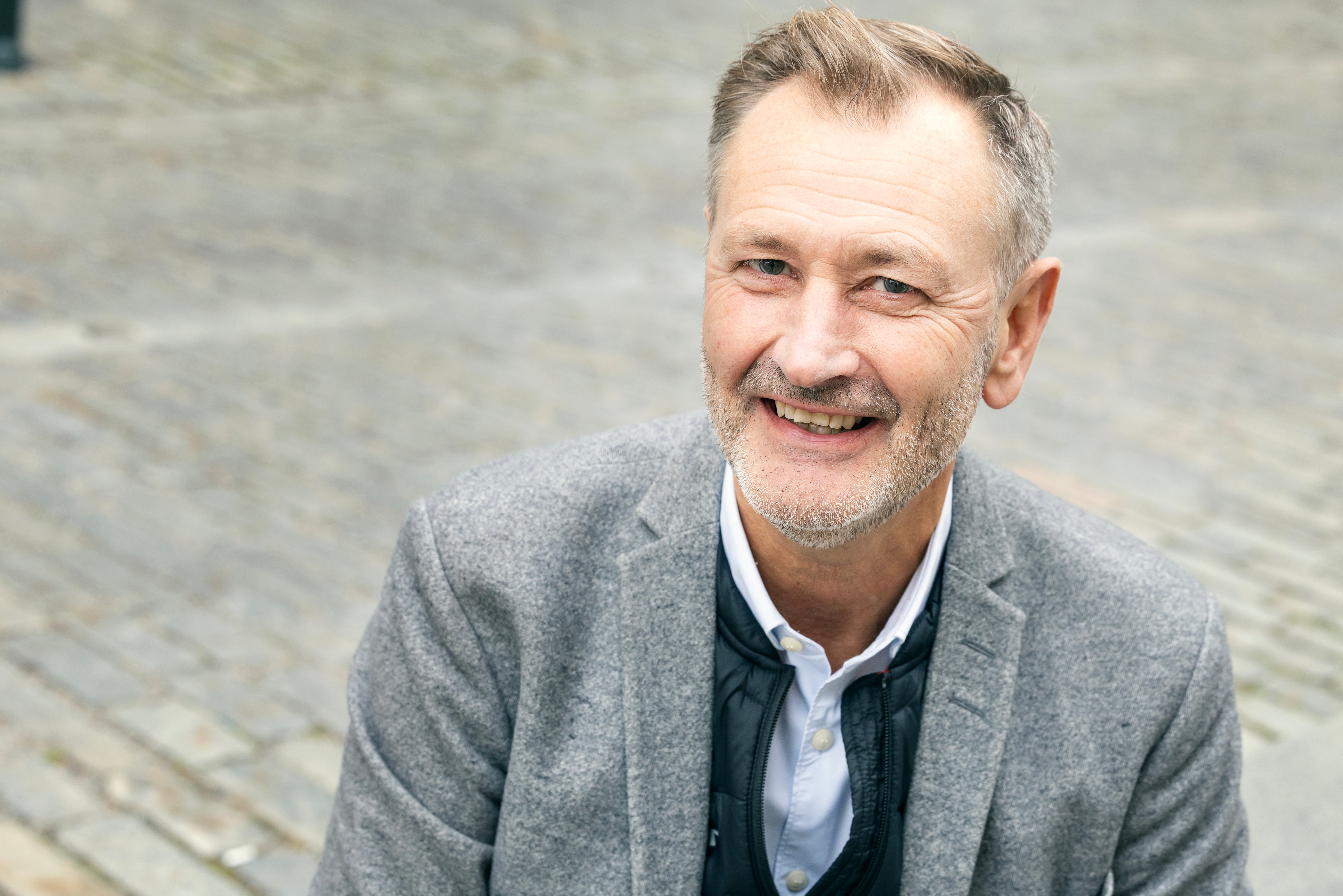 En leende man i grå kavaj sitter utomhus på en gata med gatsten.