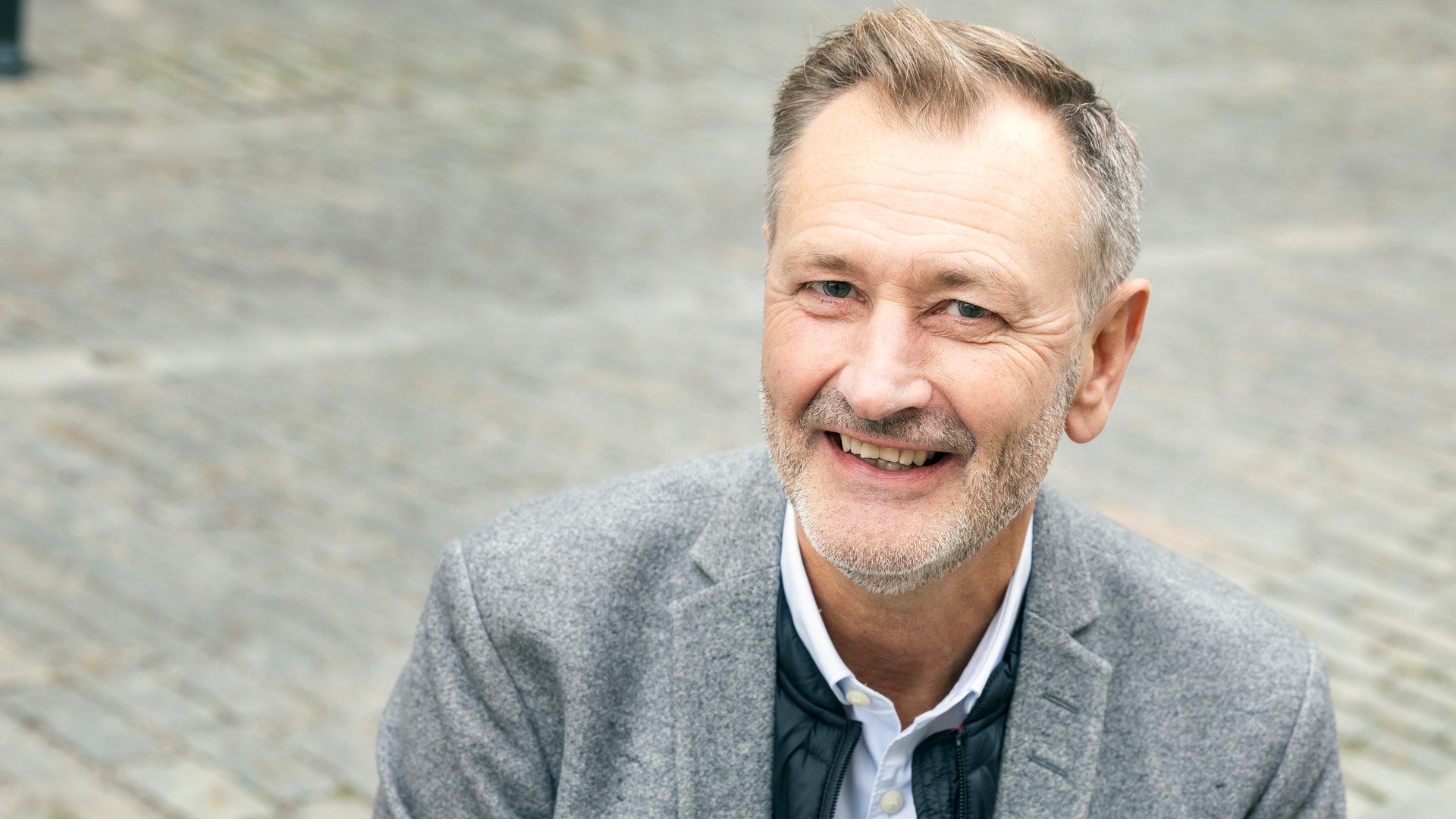 En leende man i grå kavaj sitter utomhus på en gata med gatsten.