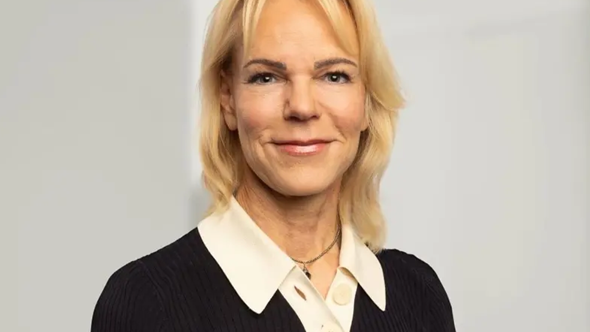 Porträtt av en kvinna med blont hår, svart jacka och vit krage som ler mot kameran.