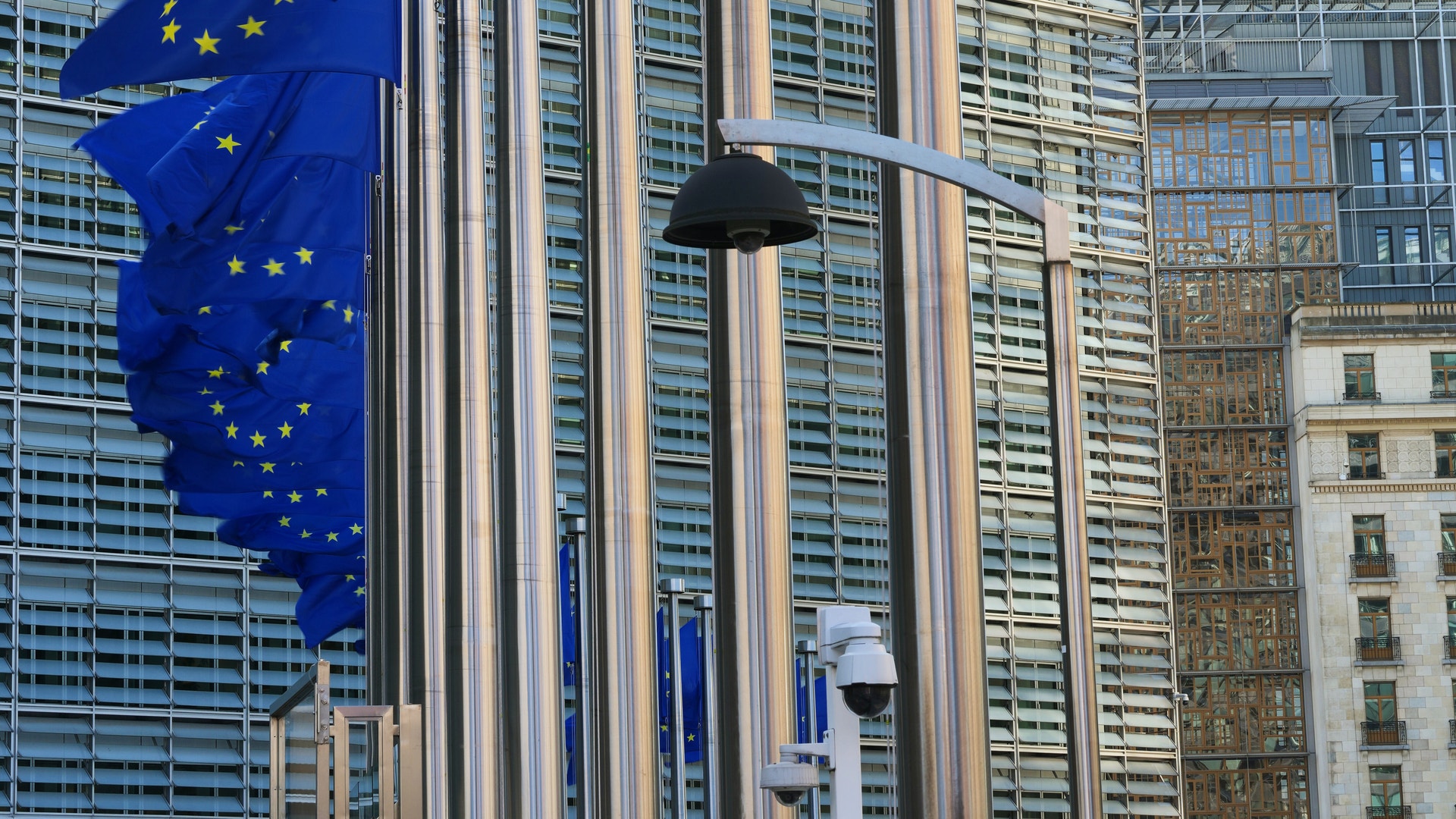 Tre EU-flaggor med blå bakgrund och gula stjärnor som hänger framför moderna kontorsbyggnader.