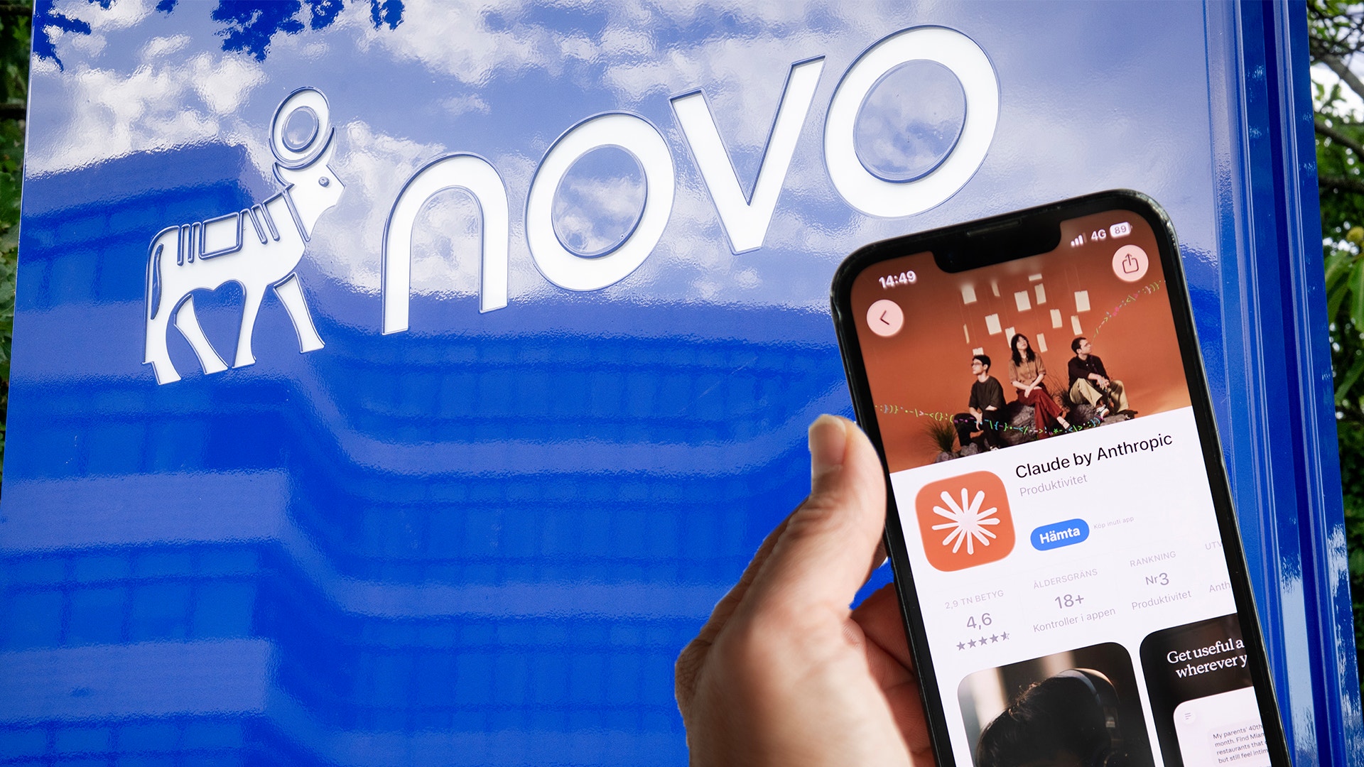 En hand håller upp en mobiltelefon med AI-appen Claude från Anthropic framför Novos logotyp på en blå skylt.