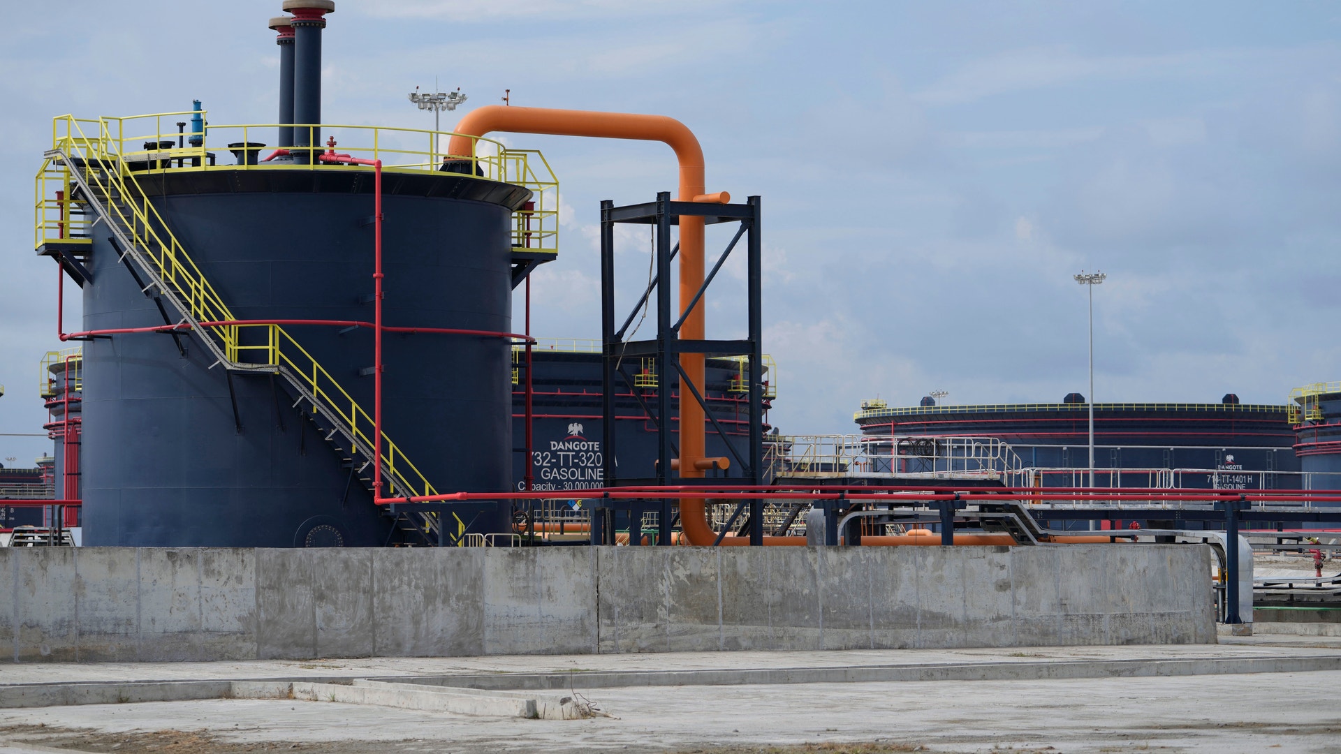 Stor industriell tank med gula säkerhetstrappor och orange rörledningar på en fabrik eller industrianläggning.