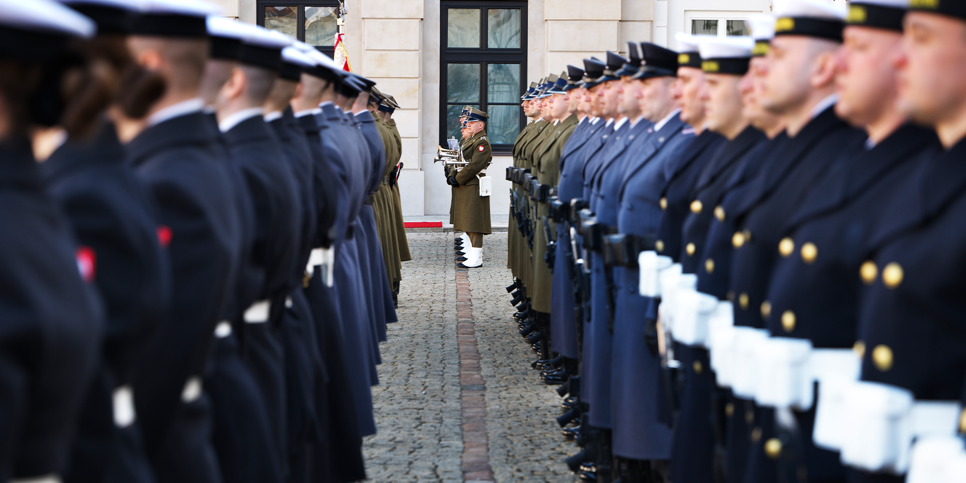 Militär ceremoni med soldater i uniform uppställda på båda sidor av en gata framför en byggnad.