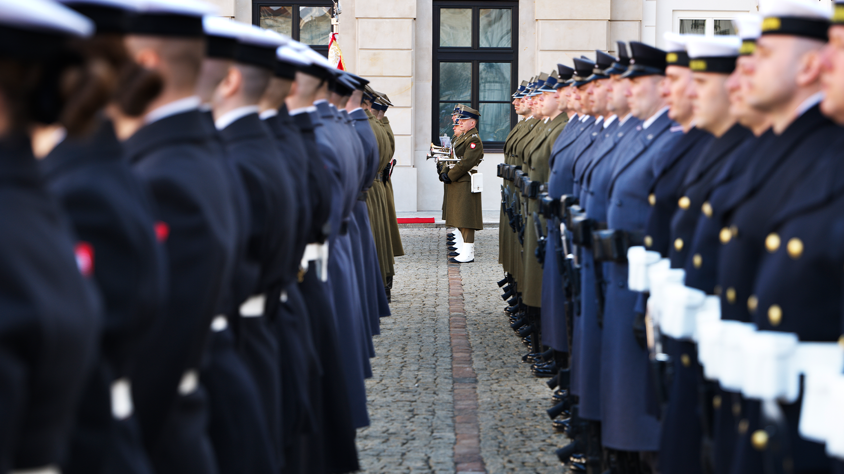 Militär ceremoni med soldater i uniform uppställda på båda sidor av en gata framför en byggnad.