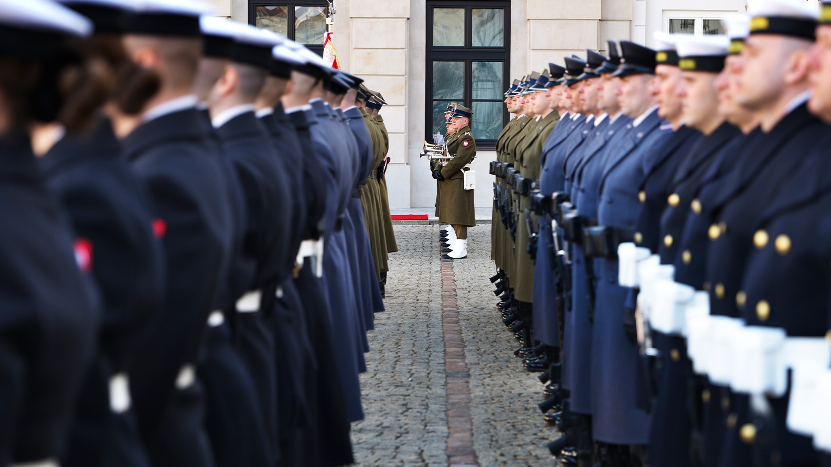 Militär ceremoni med soldater i uniform uppställda på båda sidor av en gata framför en byggnad.