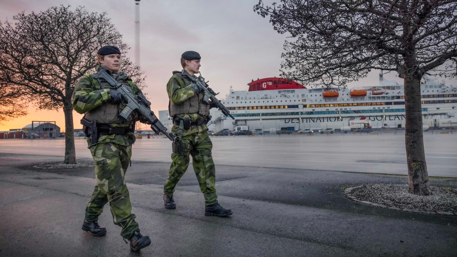 Två soldater i kamouflageuniformer patrullerar vid en hamn. I bakgrunden ligger en stor färja vid kajen under gryningen.