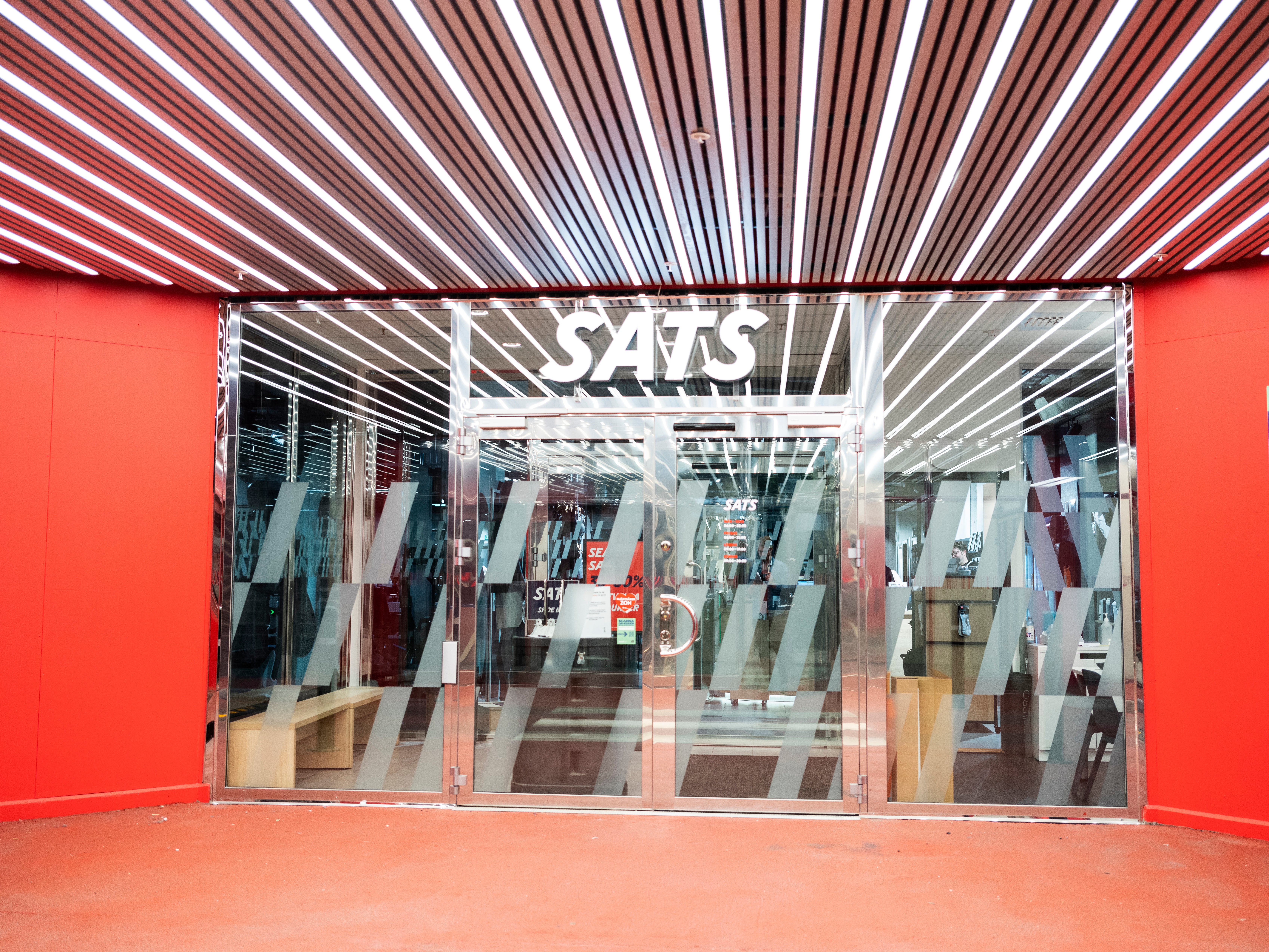 Butiksfasad med röda väggar och glasdörrar. Inuti syns kläder hängande på ställningar. Ovanför butiken finns ett vitt logotyp med texten SATS och ett modernt taklampor med röda och vita strimmor.