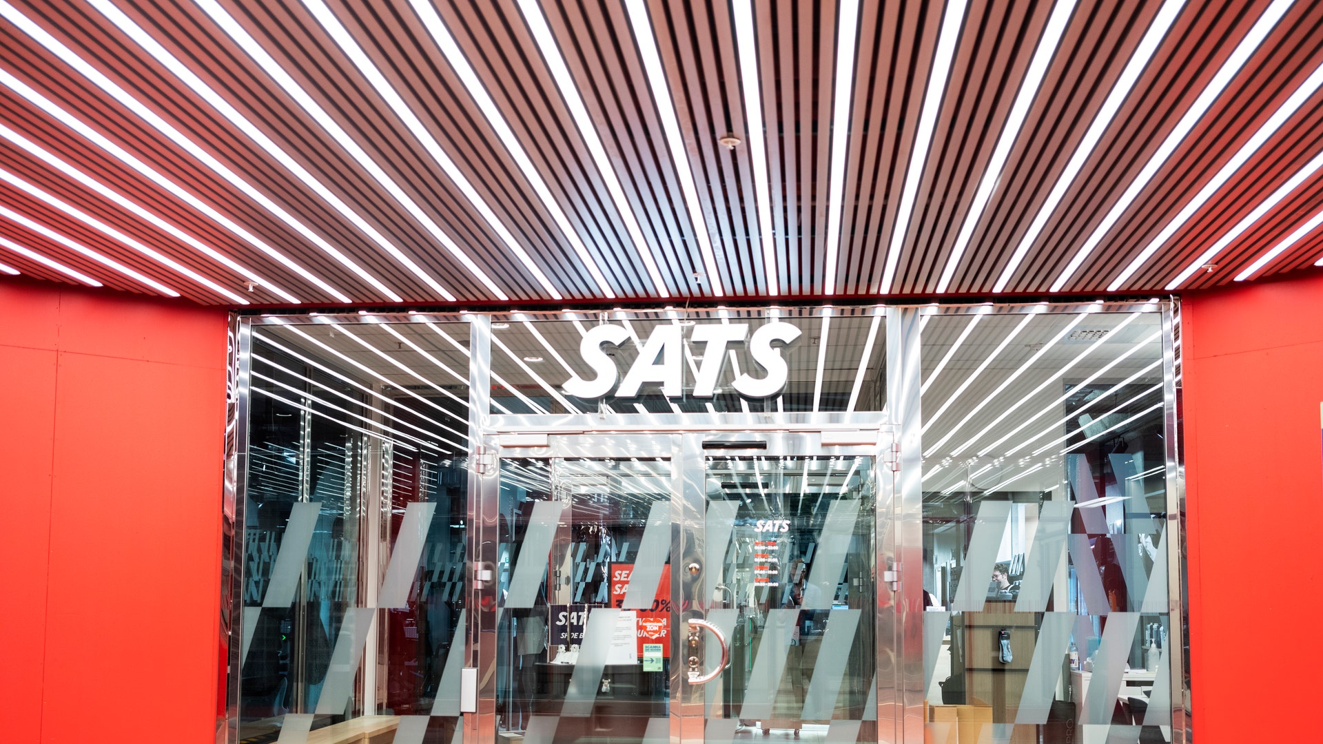 Butiksfasad med röda väggar och glasdörrar. Inuti syns kläder hängande på ställningar. Ovanför butiken finns ett vitt logotyp med texten SATS och ett modernt taklampor med röda och vita strimmor.