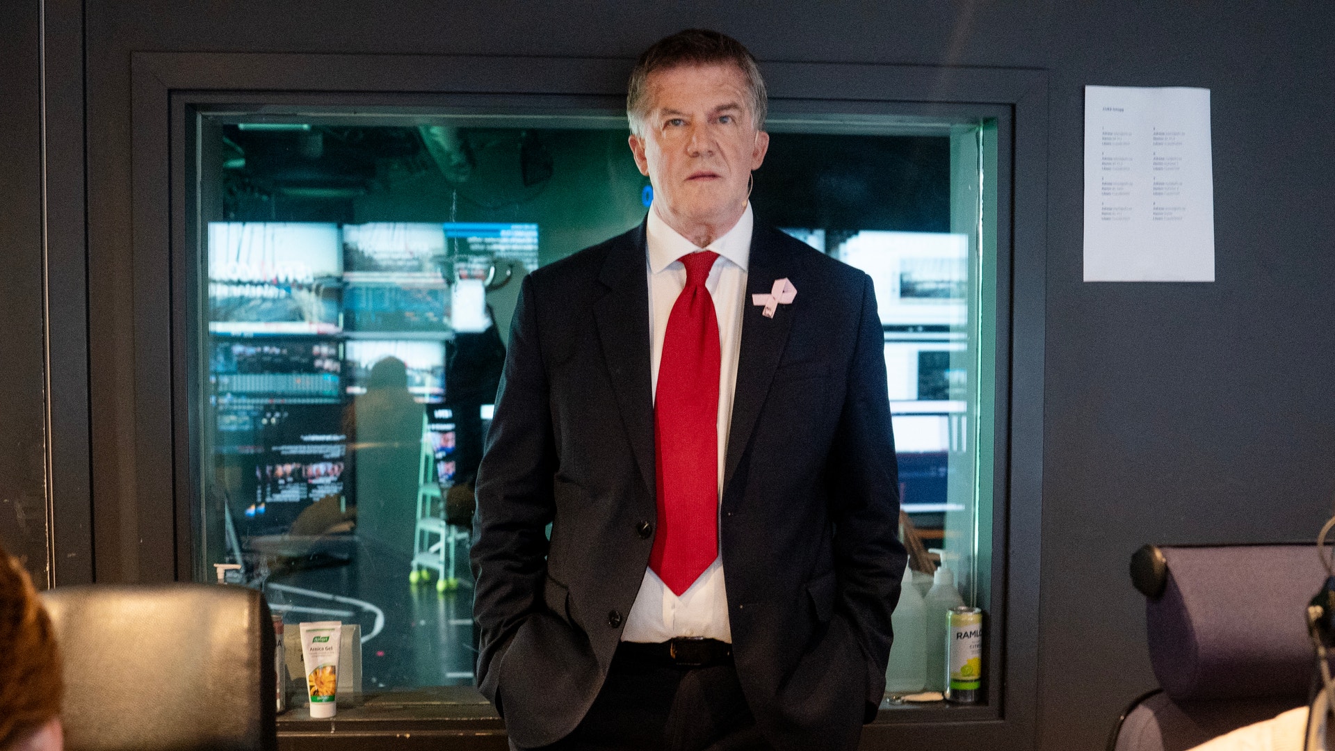 Ilja Batljan i mörk kostym och röd slips står med händerna i fickorna framför ett fönster in till en studio.