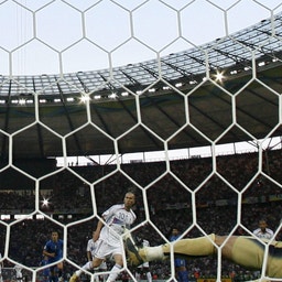 En fotbollsmatch där en boll är på väg in i målet. Bilden är tagen från bakom målnätet med målvakten i gult som försöker rädda på en stor stadion.