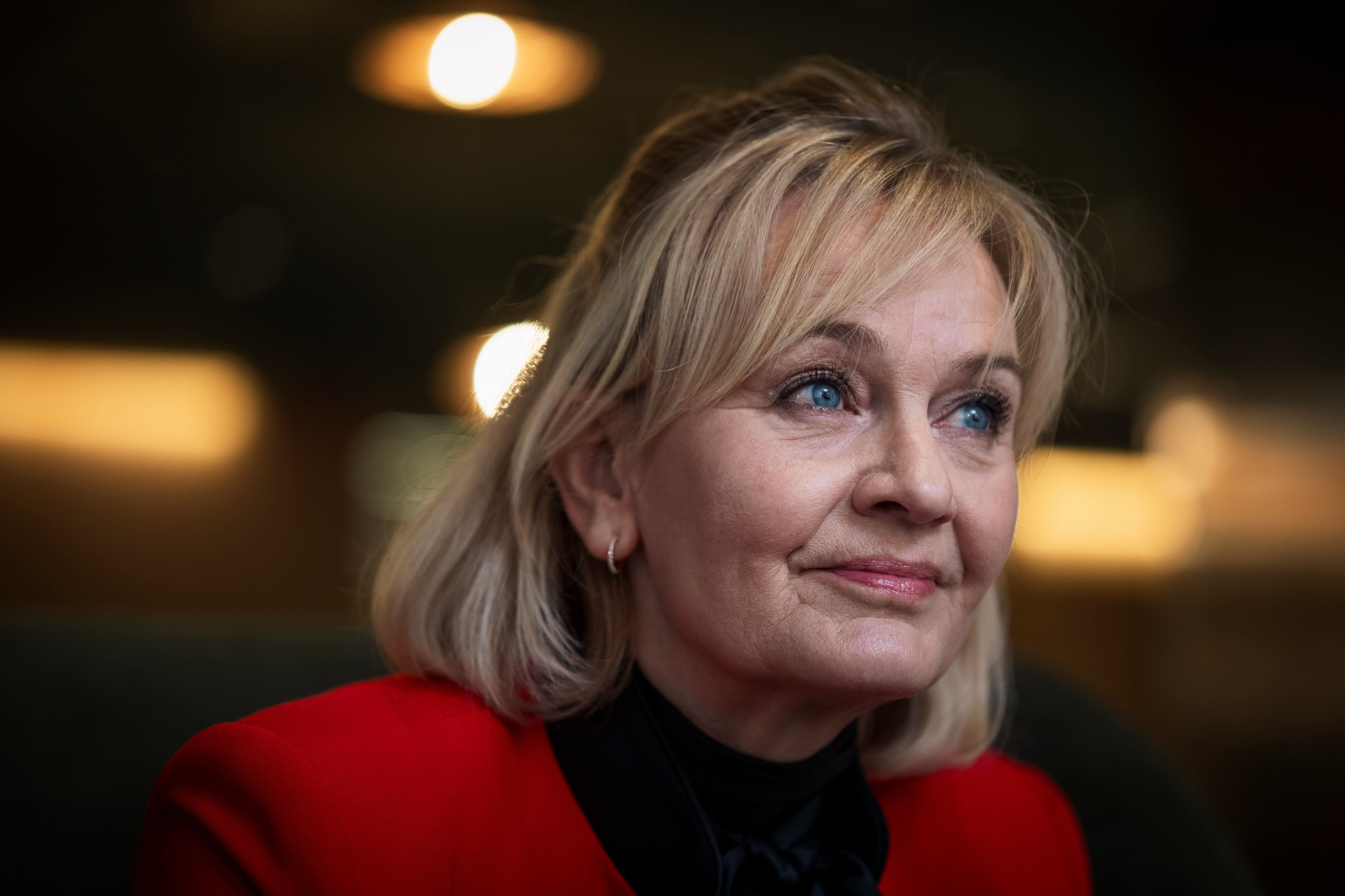 Porträttfoto av en äldre kvinna med kort ljust hår, klädd i röd jacka och svart tröja, mot en suddig bakgrund med varma ljus.
