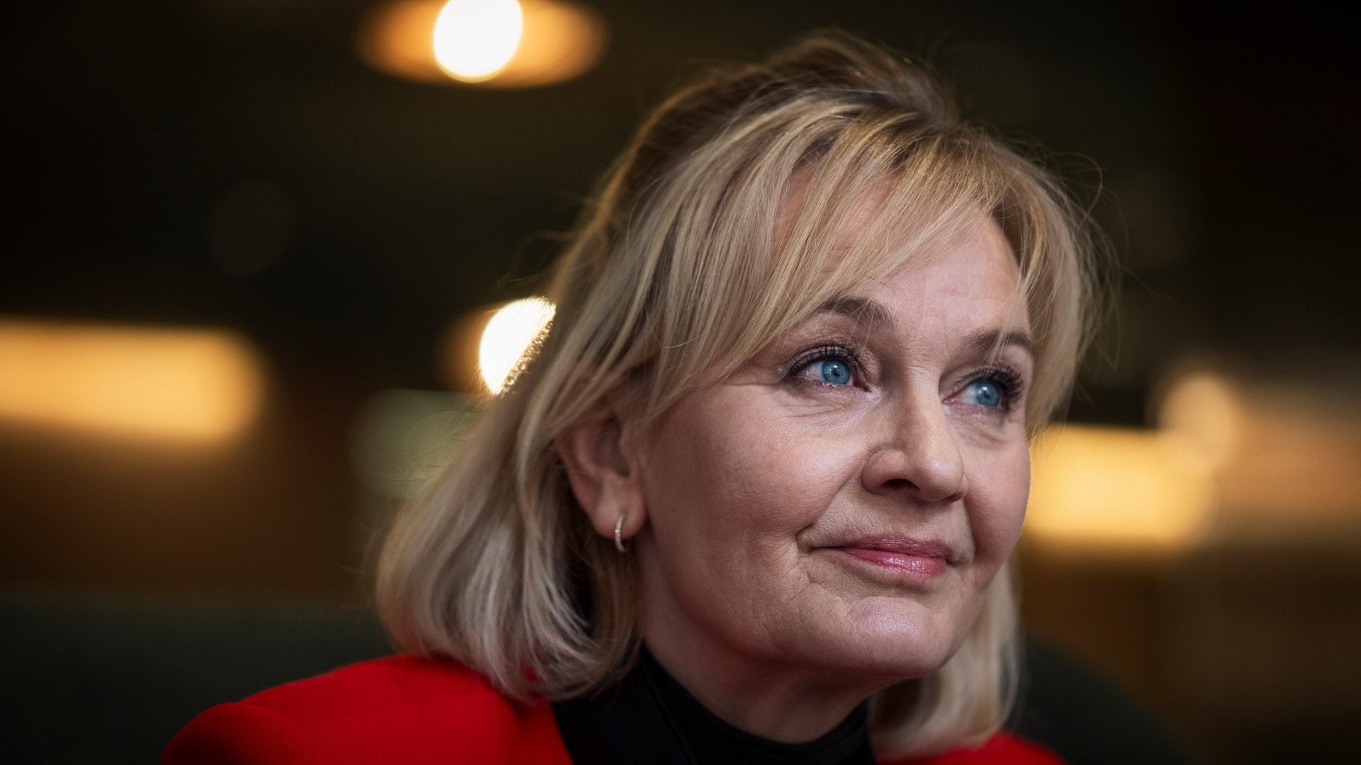 Porträttfoto av en äldre kvinna med kort ljust hår, klädd i röd jacka och svart tröja, mot en suddig bakgrund med varma ljus.