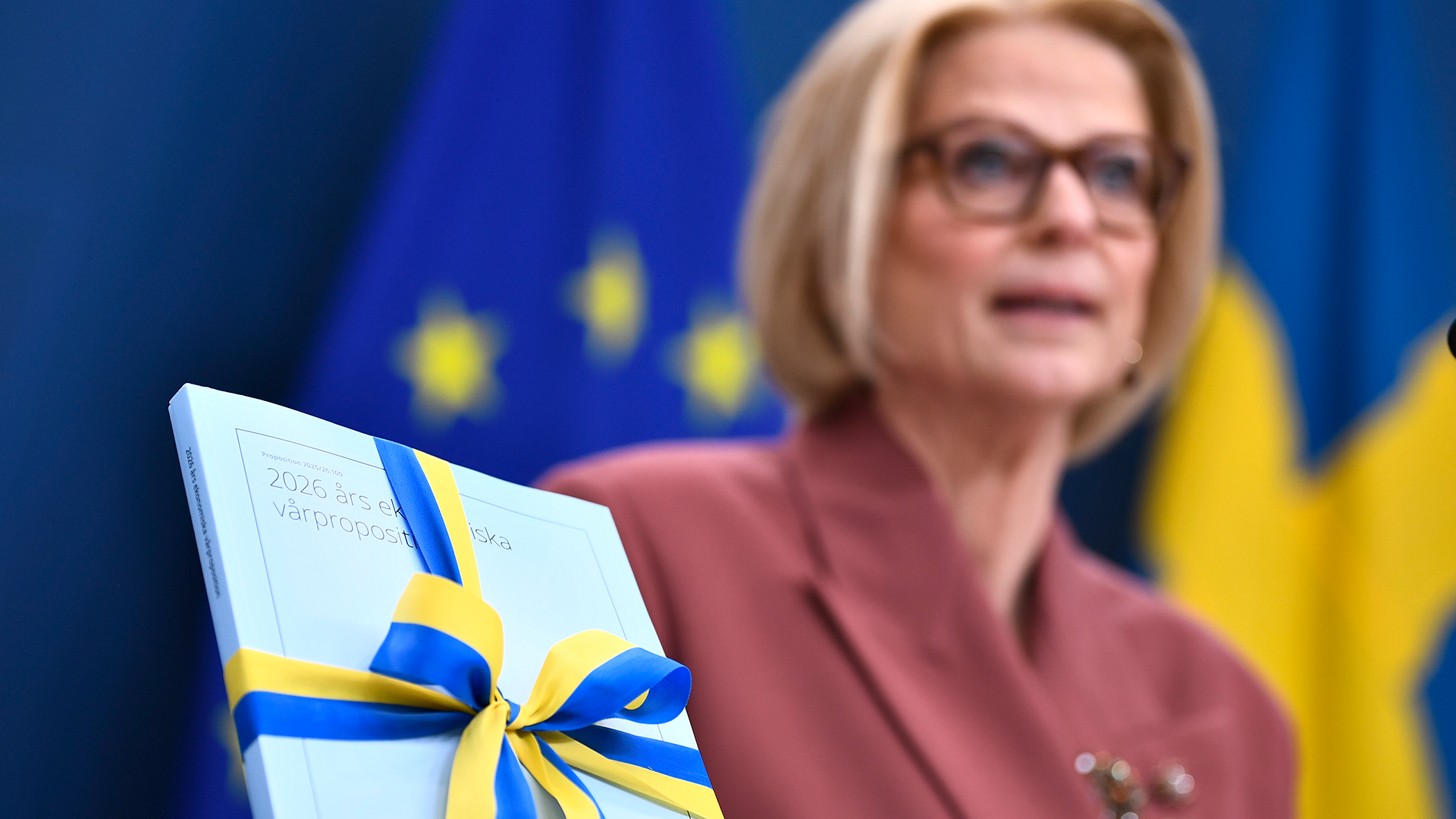 En kvinna i röd jacka bakom en presentask inslöjad med blå och gula band, med EU och svenska flaggor i bakgrunden.