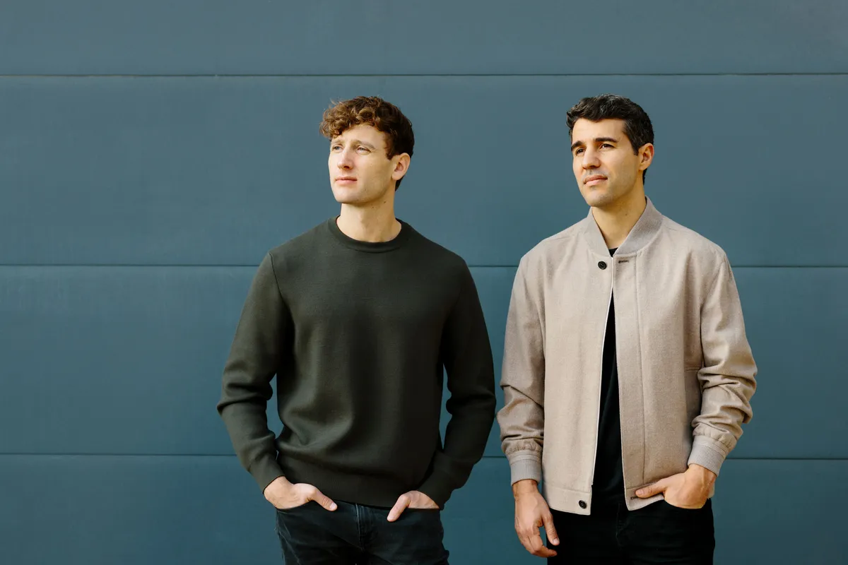 Två män står framför en blå vägg. Den vänstra mannen bär en mörkgrön tröja och den högra bär en beige jacka över en svart skjorta.