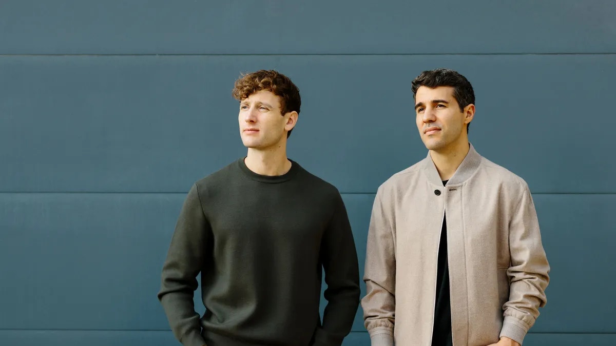 Två män står framför en blå vägg. Den vänstra mannen bär en mörkgrön tröja och den högra bär en beige jacka över en svart skjorta.