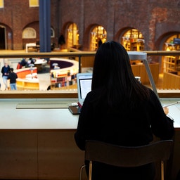 Två personer sitter vid ett högt bord med laptops och tittar ut genom ett stort fönster. Bakom fönstret syns en vacker arkitektonisk miljö med en tegelbyggnad och människor.