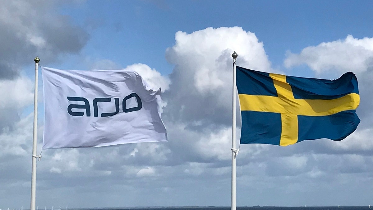 Två flaggor som vajar mot en blå himmel med moln. Till vänster ARJO-flaggan och till höger den svenska flaggan.