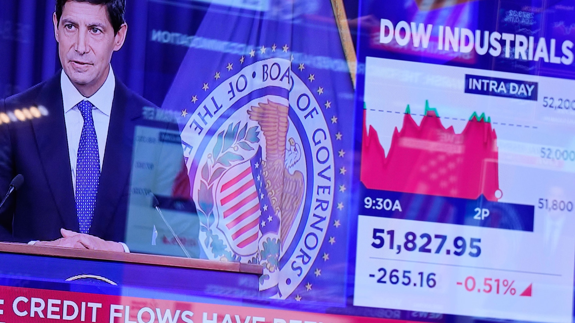 En man håller pressanförande vid en talarstol med Federal Reserves emblem, bredvid en skärm som visar börskurser för Dow Jones industriindex.
