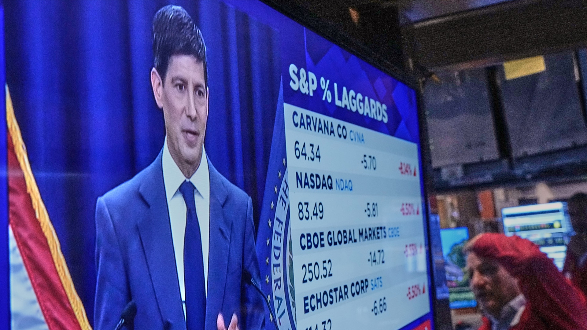 En tv-skärm på en börshandelsgolv visar en man i kostym som talar, tillsammans med en lista över aktiekurser.